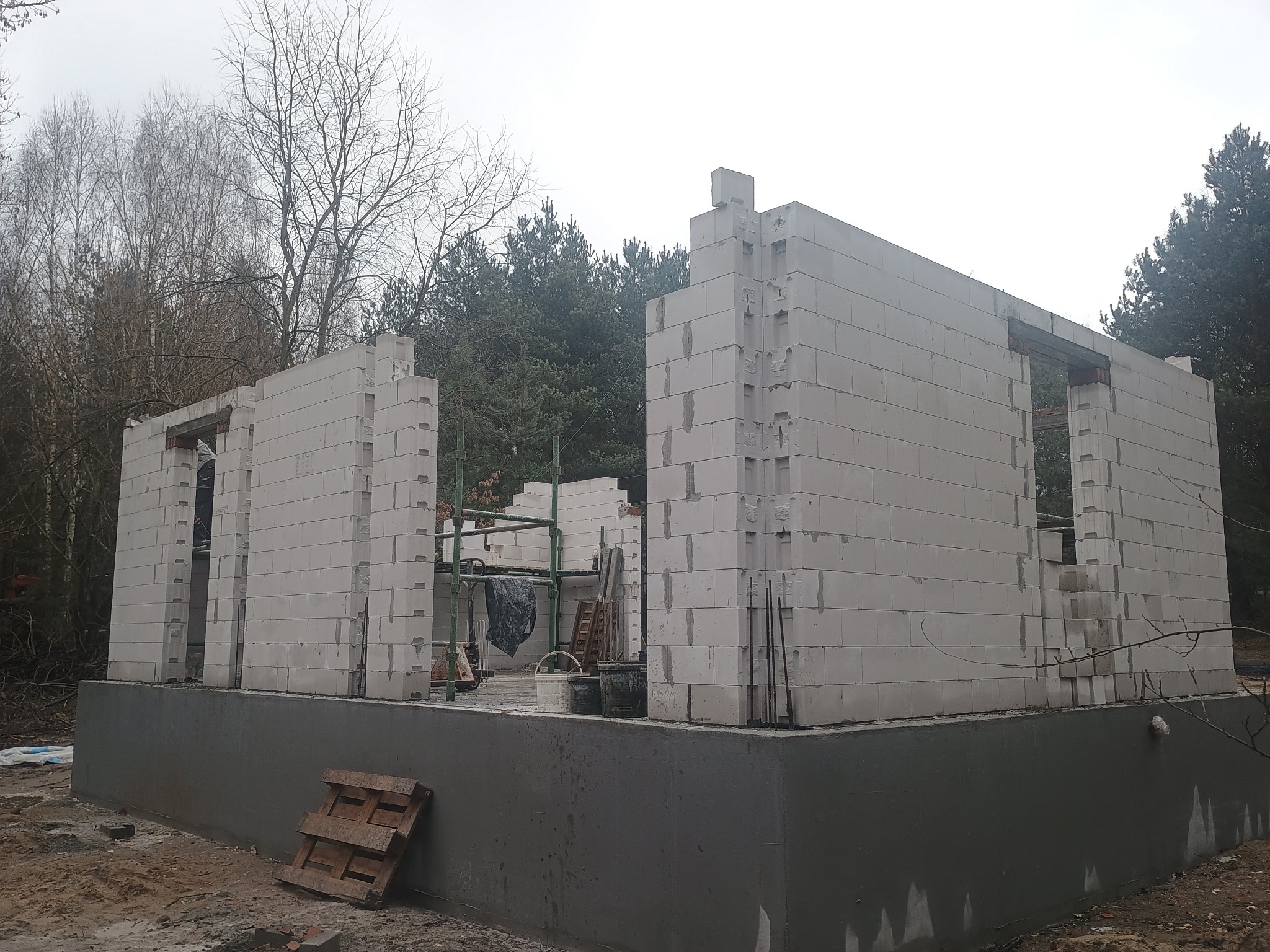 Wznoszenie ścian domu z betonu komórkowego na fundamencie. Widoczne rusztowania i materiały budowlane. W tle las i pochmurne niebo.