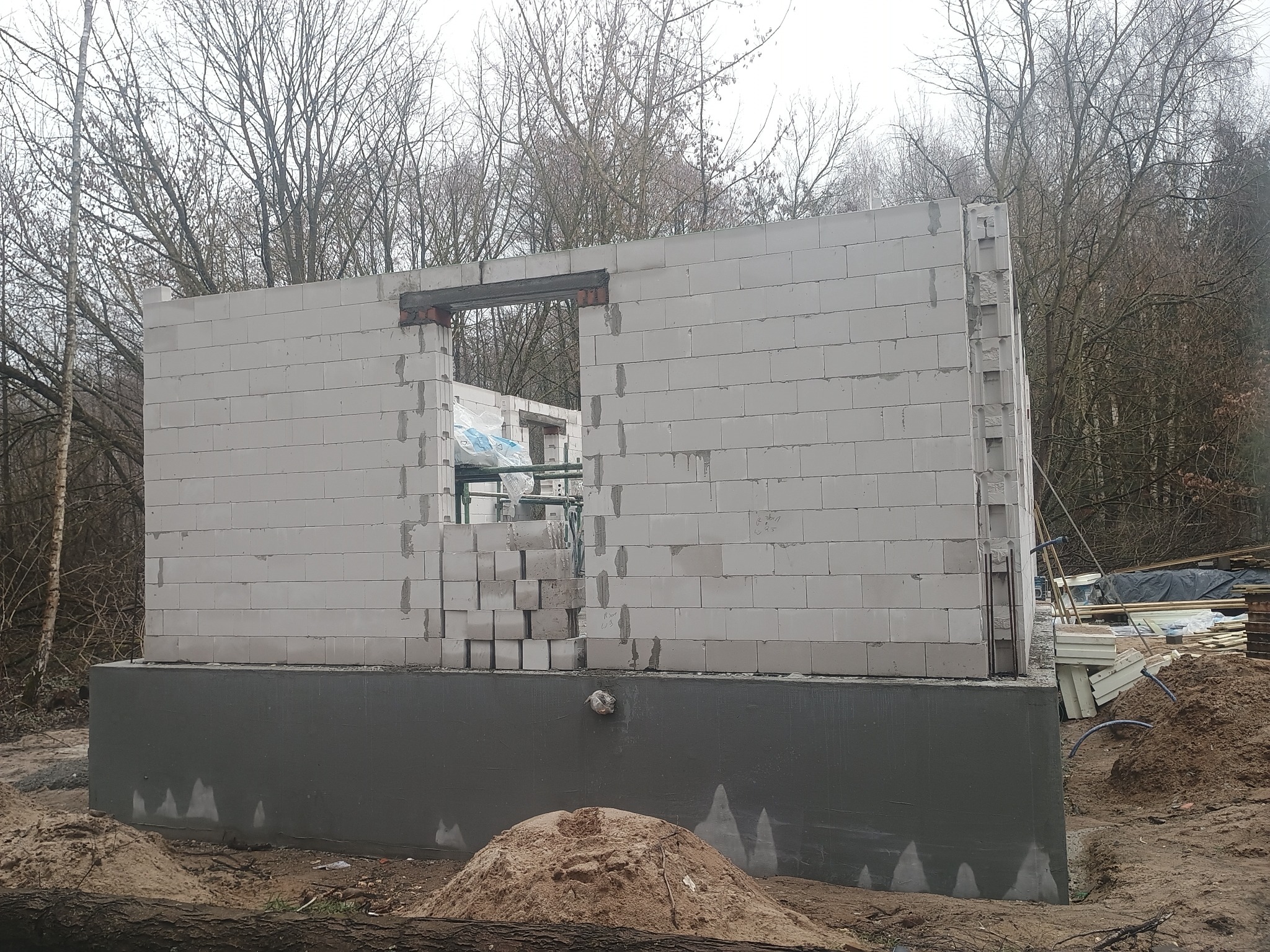Wznoszenie ścian parteru z betonu komórkowego na fundamencie. Widoczne zbrojenie nad otworem okiennym. W tle drzewa bez liści i plac budowy.