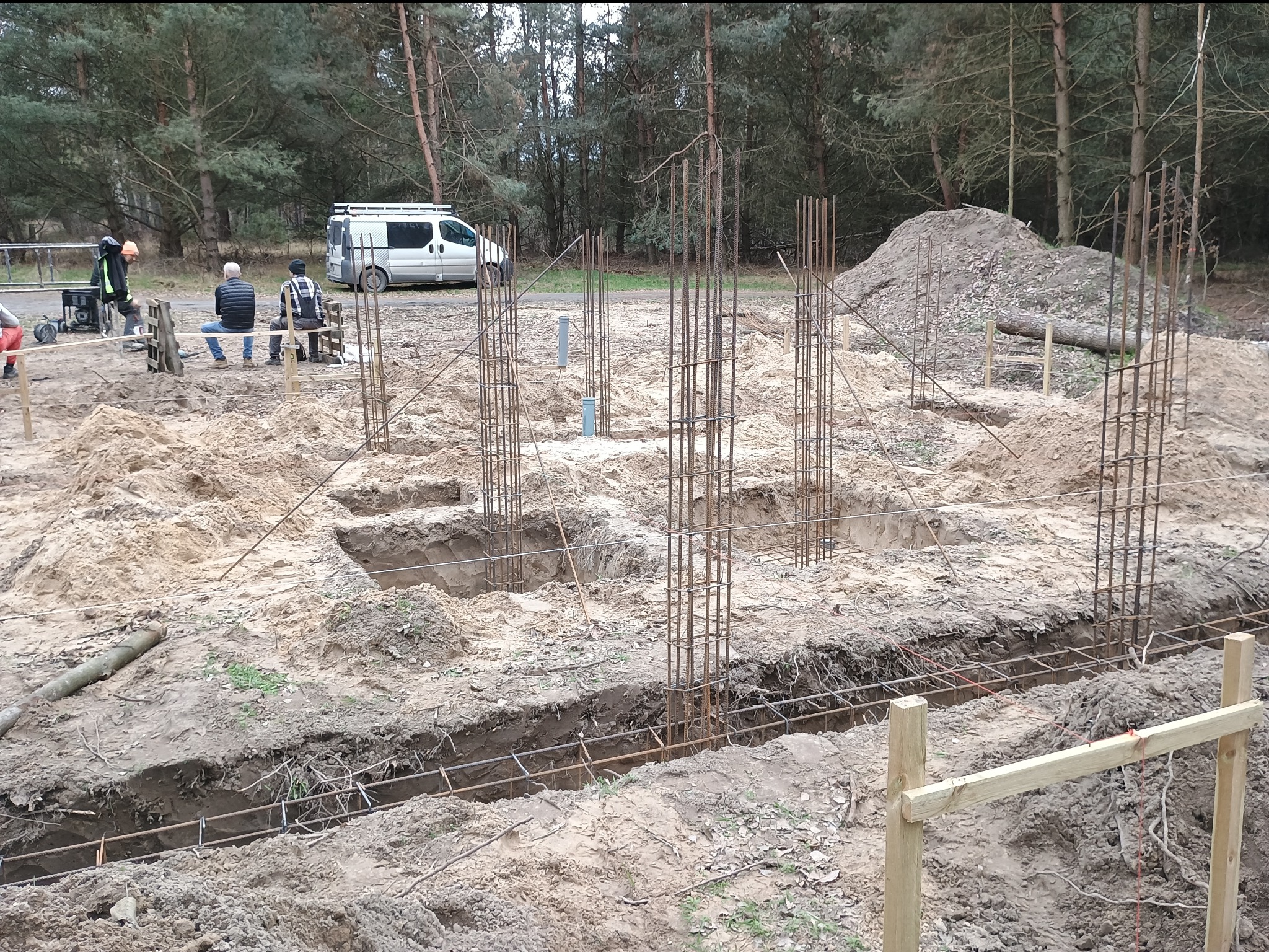 Przygotowanie fundamentów pod budowę domu: wykopy, zbrojenie stalowe, szalunki w ziemi. W tle ekipa budowlana i samochód dostawczy na skraju lasu.