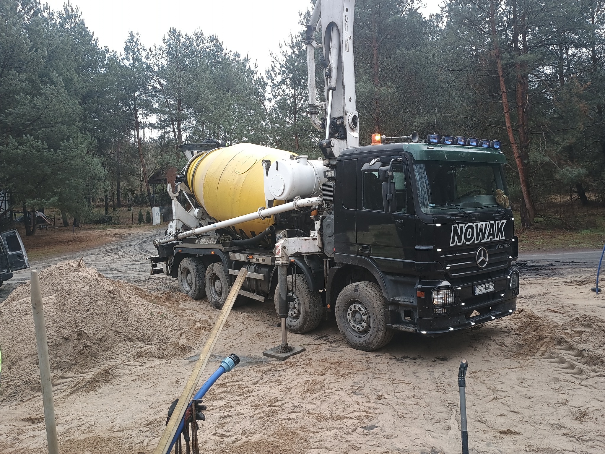 Ciężarówka z betonomieszarką na placu budowy, z widoczną stertą piasku i sprzętem. W tle las iglasty.