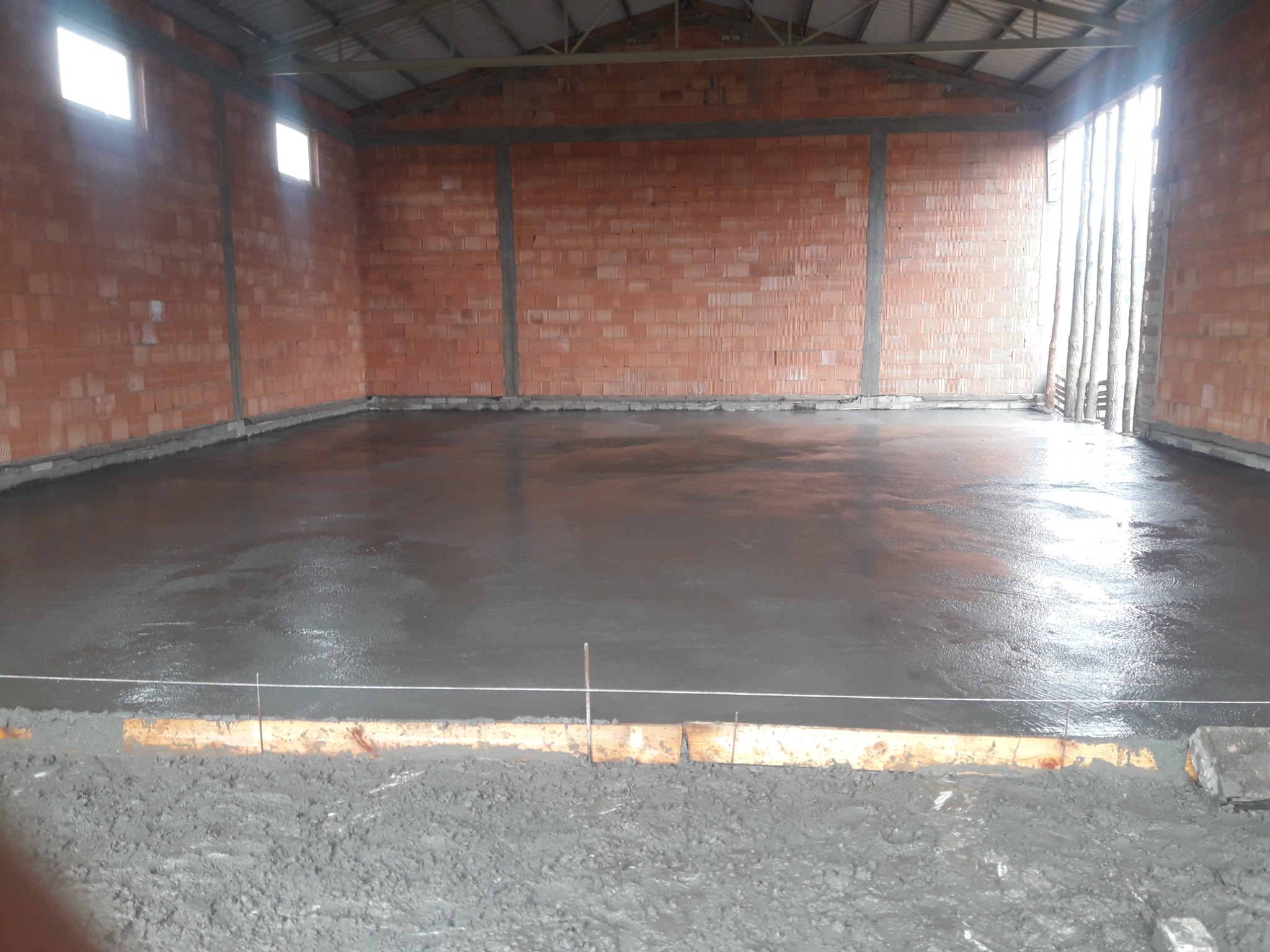 Wnętrze surowego domu w trakcie budowy: ceglane ściany, wylana, mokra posadzka z widocznymi sznurami poziomującymi i deskami szalunkowymi. Widoczne okna i konstrukcja dachu.