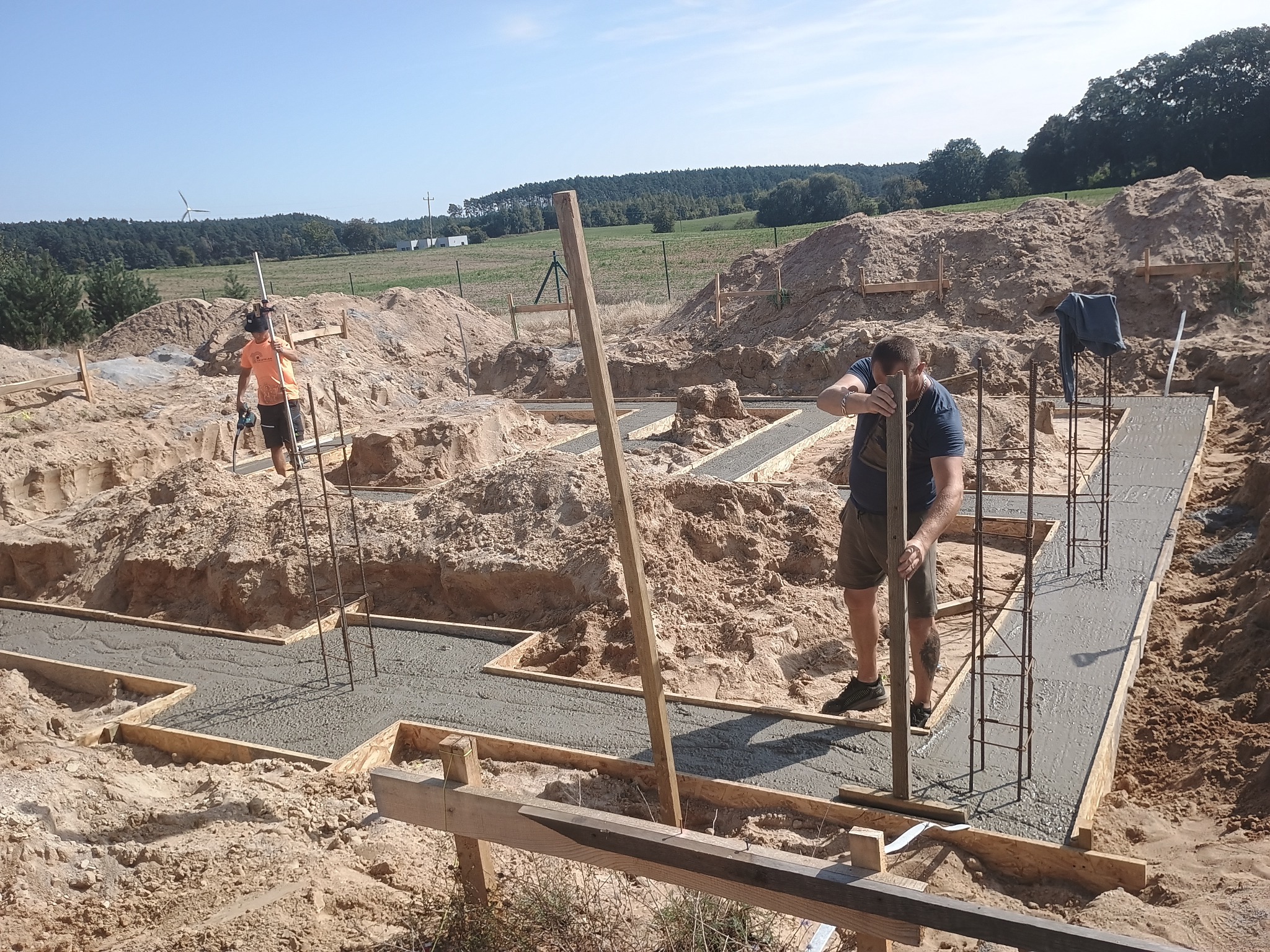 Wylewanie fundamentów pod dom na piaszczystym terenie. Widoczne drewniane szalunki i stalowe zbrojenia. Dwóch pracowników budowlanych w trakcie pracy.