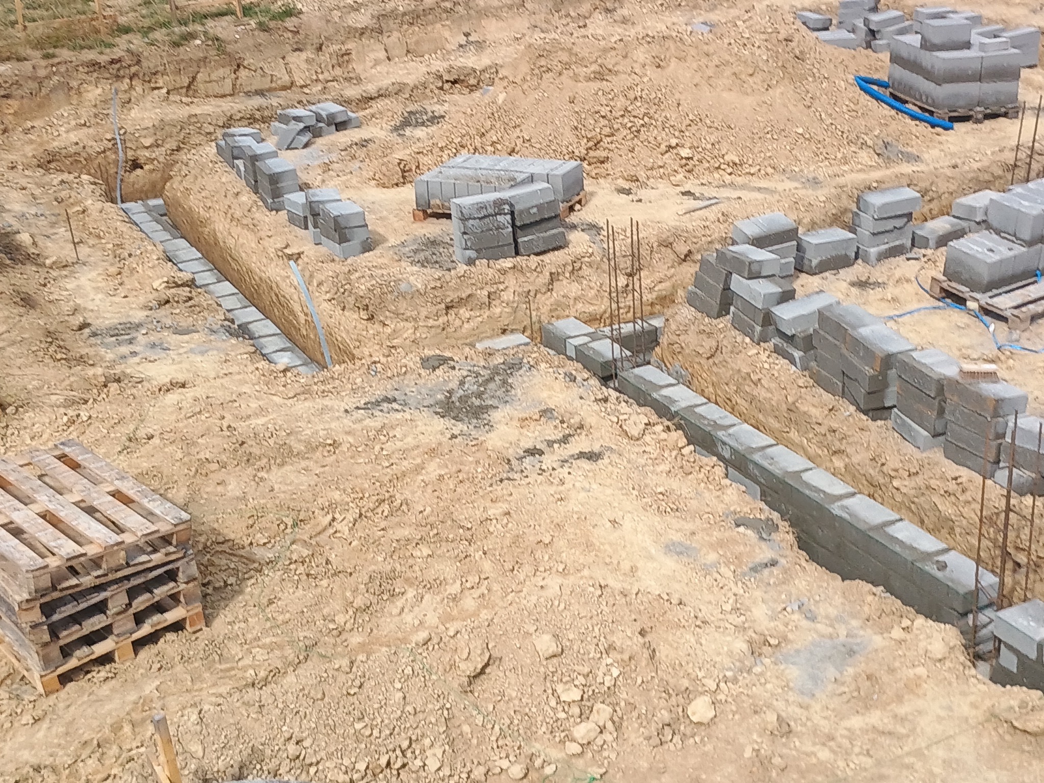 Fundamenty domu w trakcie budowy: wykopane rowy, częściowo wylane fundamenty z betonowych bloczków, zbrojenie. Widoczne palety z bloczkami i piasek.