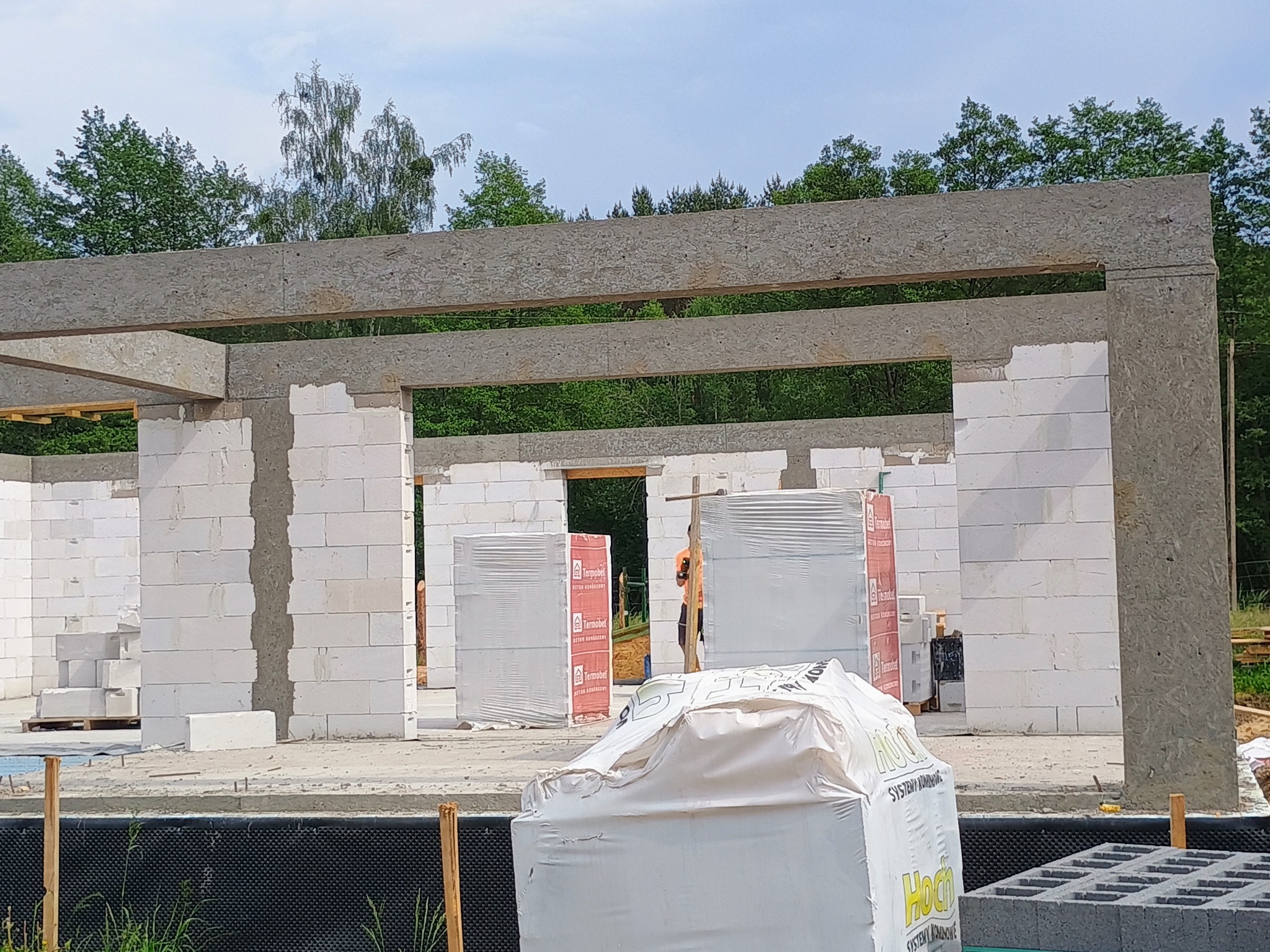 Budowa domu w trakcie: białe bloczki, betonowe nadproża, materiały budowlane na placu. Widoczny zarys ścian i otwory okienne. W tle zieleń drzew.