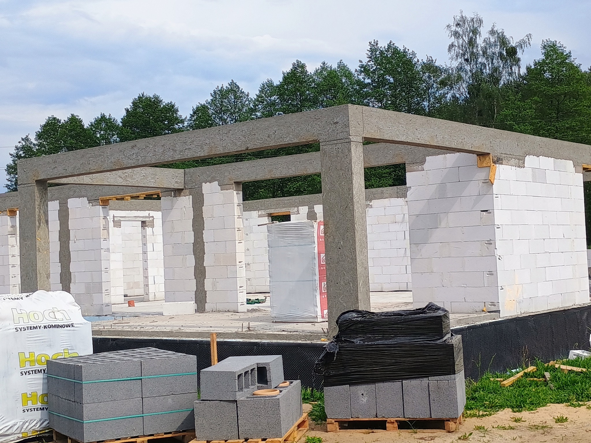 Wznoszenie domu z białych bloczków, widoczny szkielet konstrukcji i materiały budowlane. Budowa w toku, zieleń w tle.