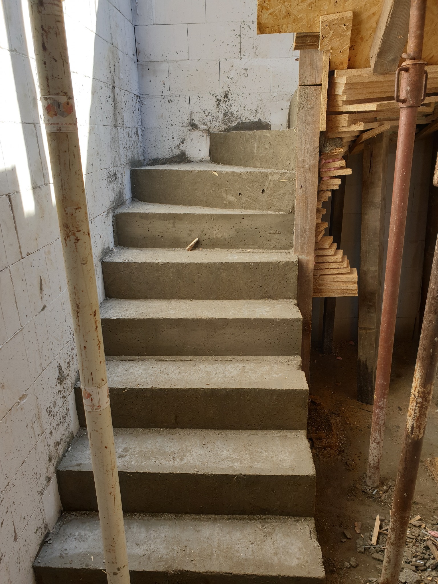 Betonowe schody w surowym stanie, widoczne podpory i szalunki. Ściany z bloczków, fragment rury podtrzymującej. Wnętrze w trakcie budowy.
