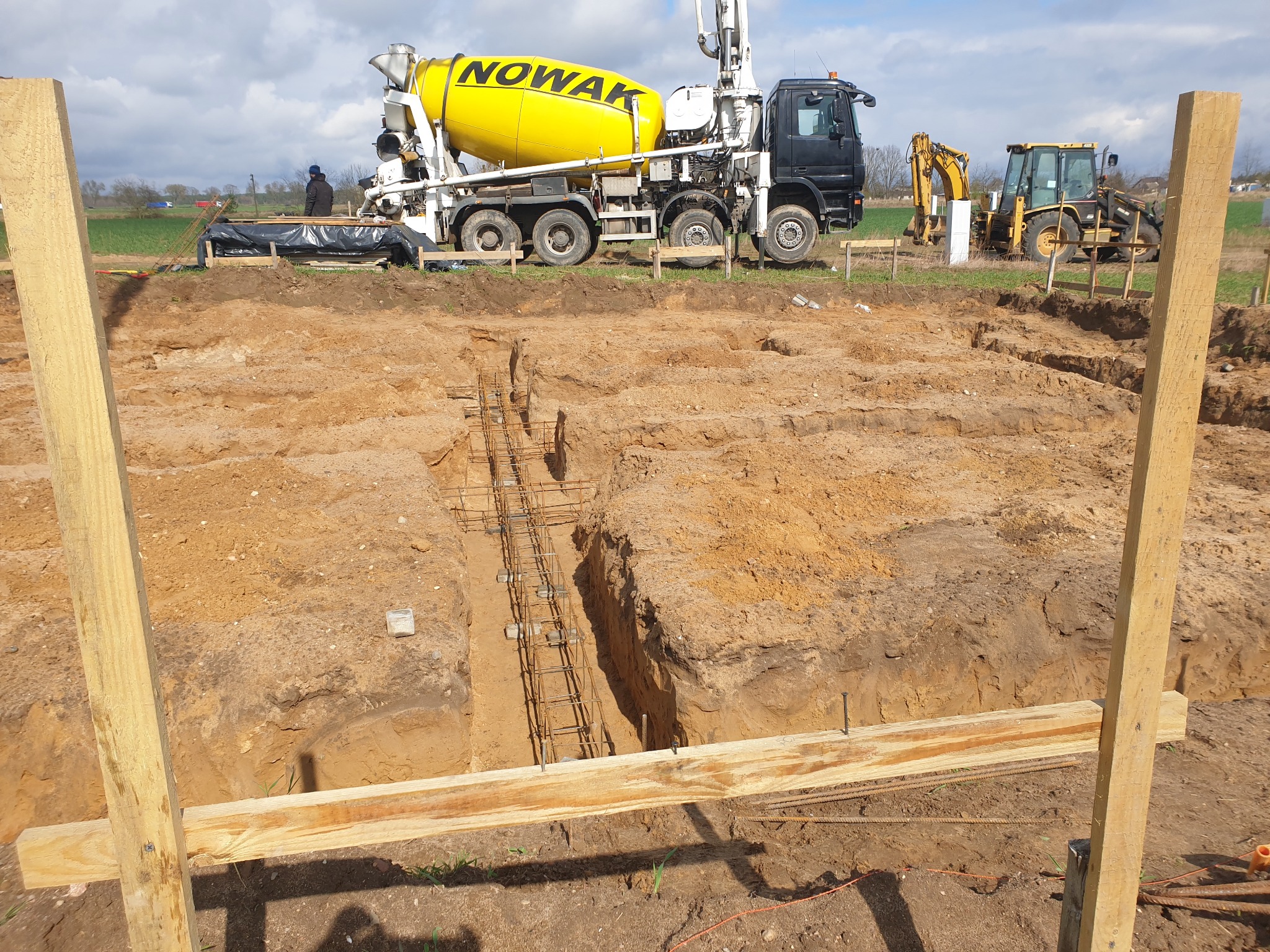 Wykop pod fundamenty z ułożonym zbrojeniem, na tle betoniarka z logo Nowak i koparki. Widok z poziomu gruntu, w słoneczny dzień.