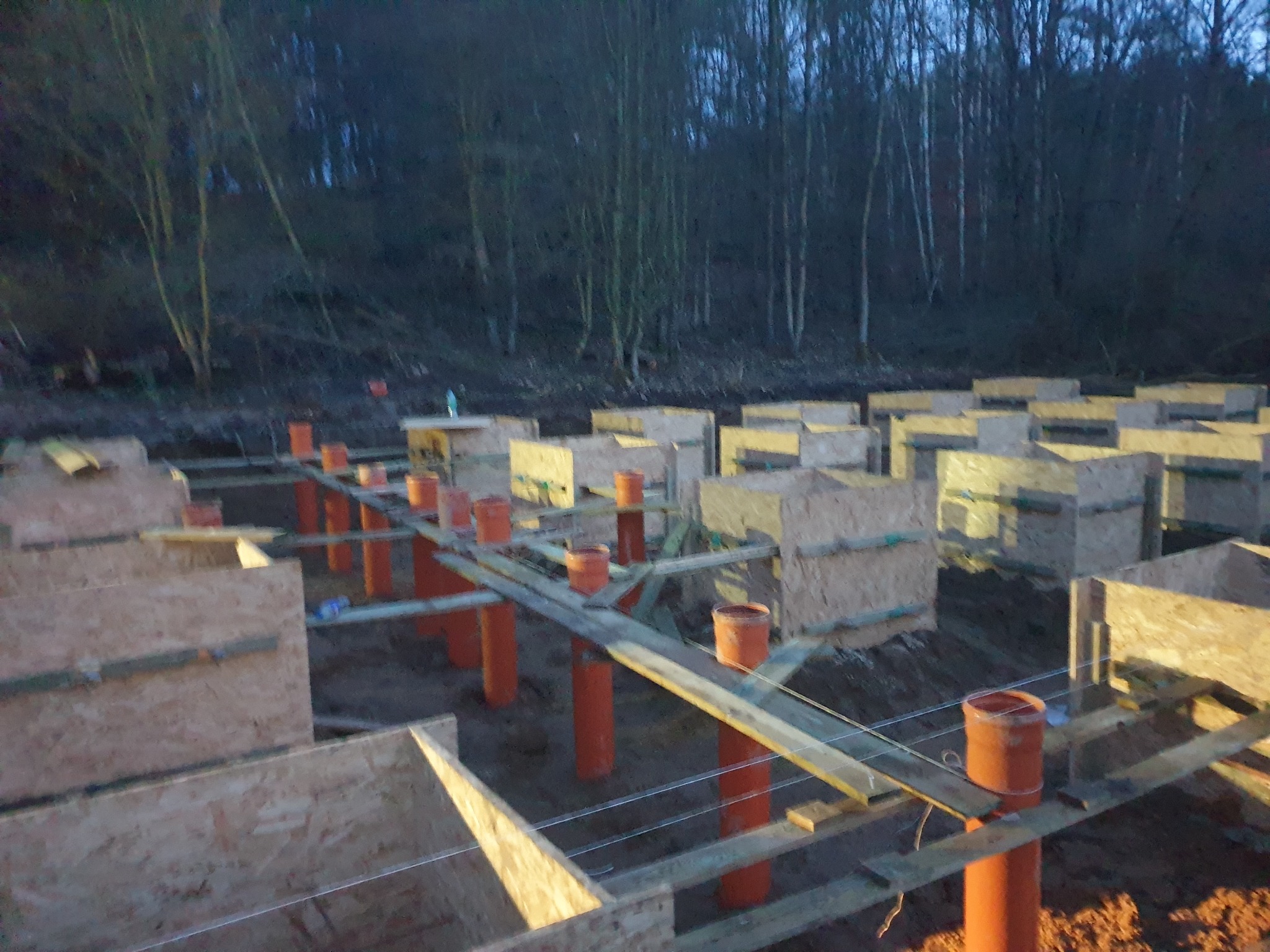 Fundamenty pod dom modułowy: drewniane szalunki i pomarańczowe rury w ziemi, przygotowane pod wylewkę. W tle drzewa.