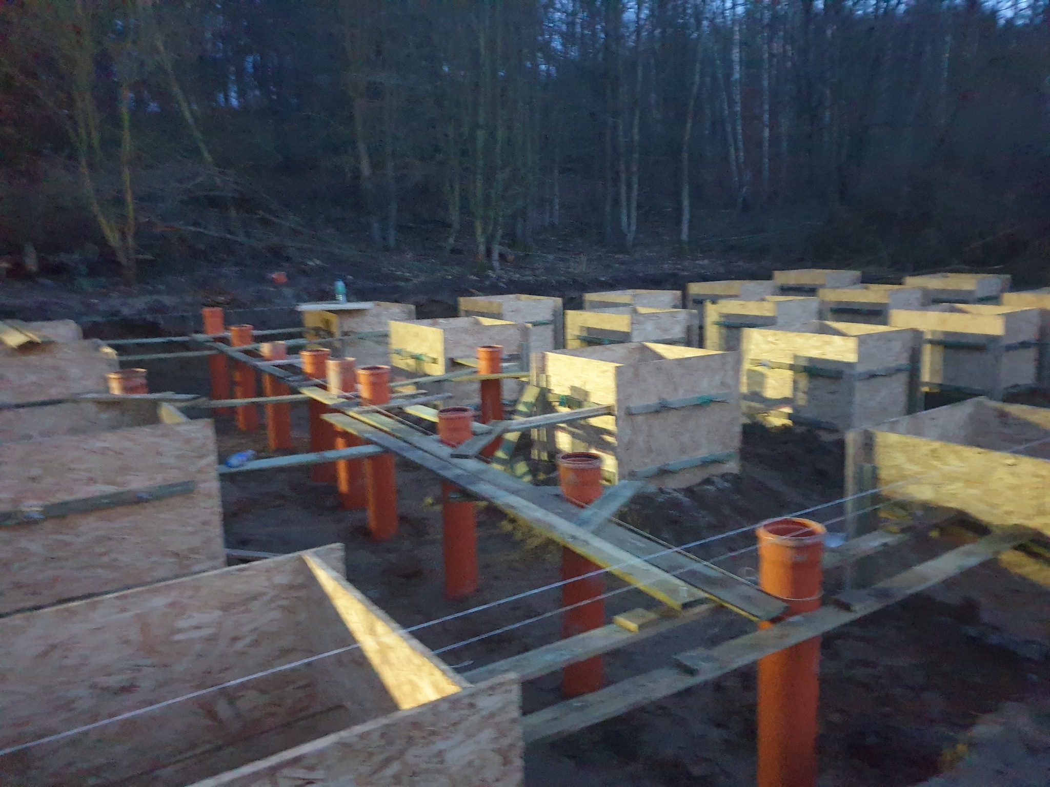 Fundamenty pod dom w trakcie budowy: drewniane szalunki i pomarańczowe rury w ziemi, widok z góry. W tle drzewa i ziemia. Etap początkowy budowy.