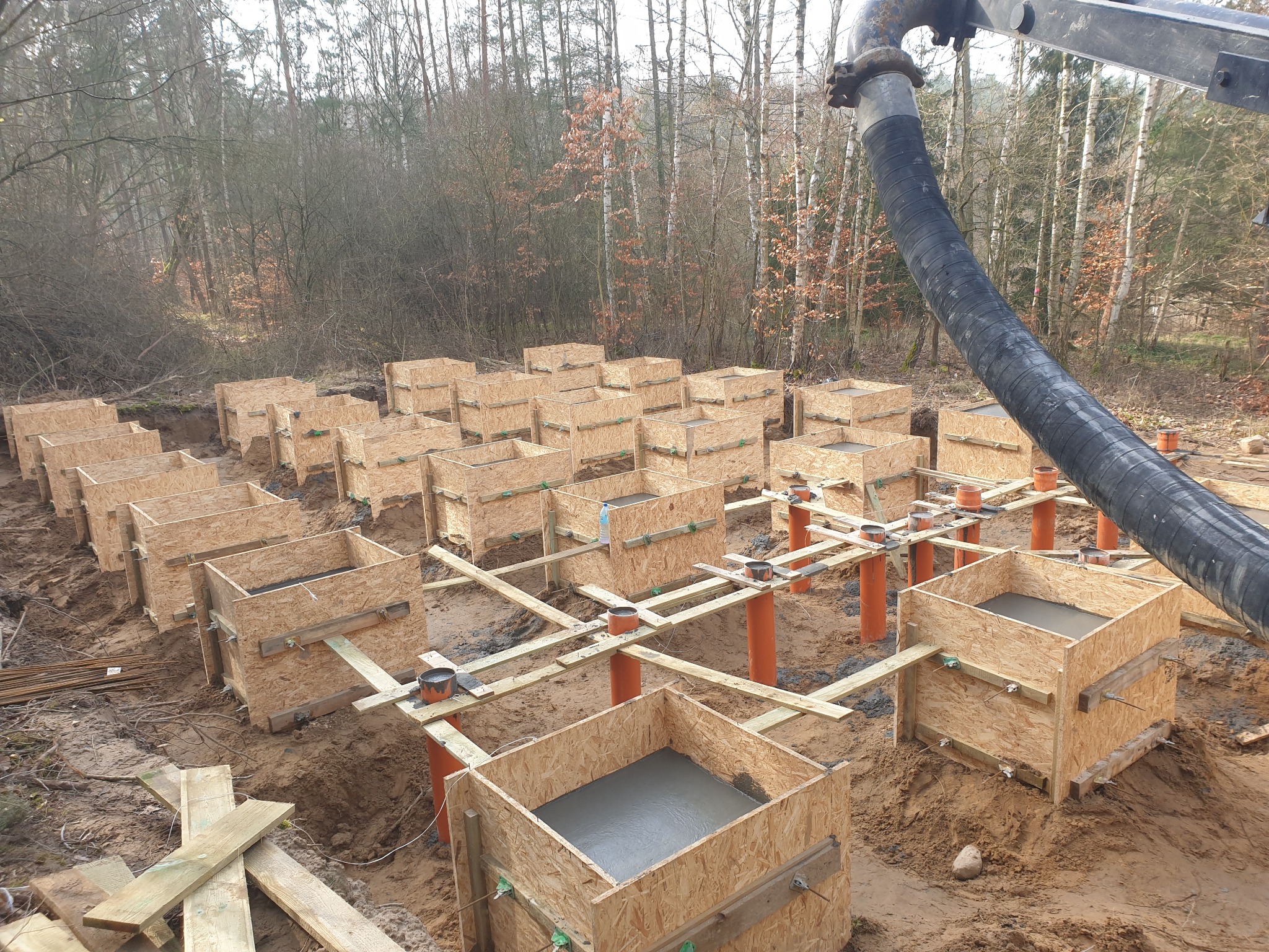 Wylane fundamenty pod dom, widoczne drewniane szalunki wypełnione betonem. W tle las, na pierwszym planie rura do pompowania betonu.