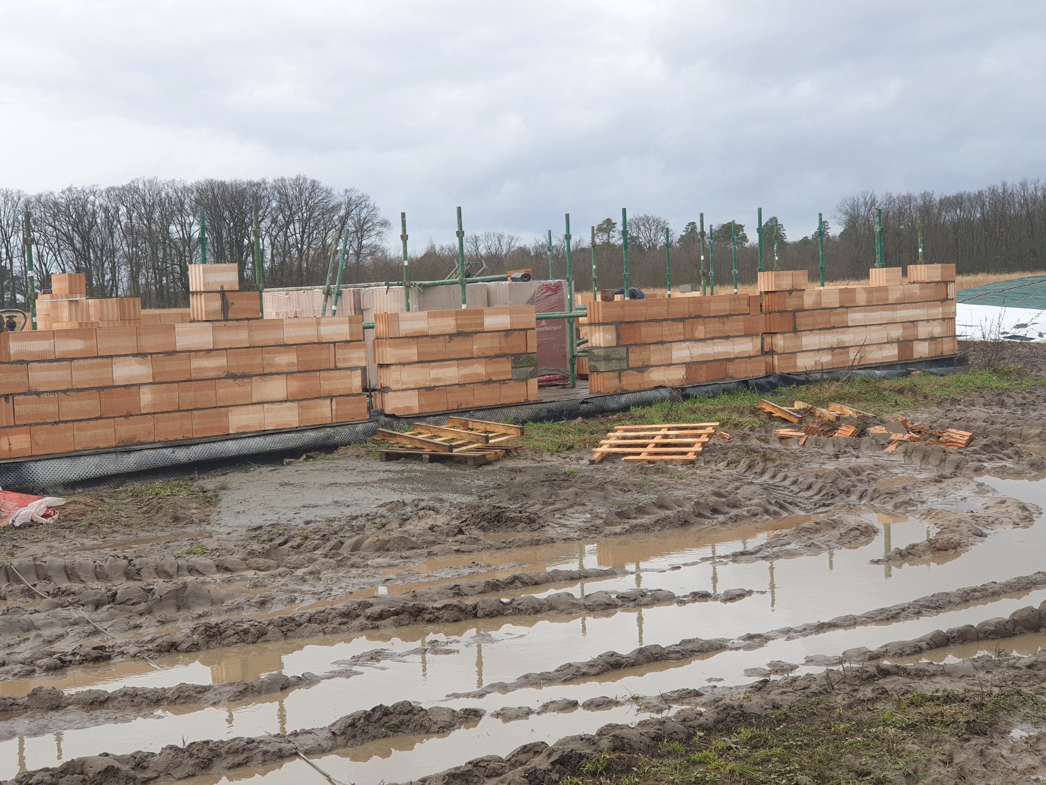 Budowa domu z czerwonej cegły ceramicznej Porotherm, widoczne ściany w trakcie wznoszenia, palety z cegłami, mokre błoto na placu budowy. Pochmurne niebo.