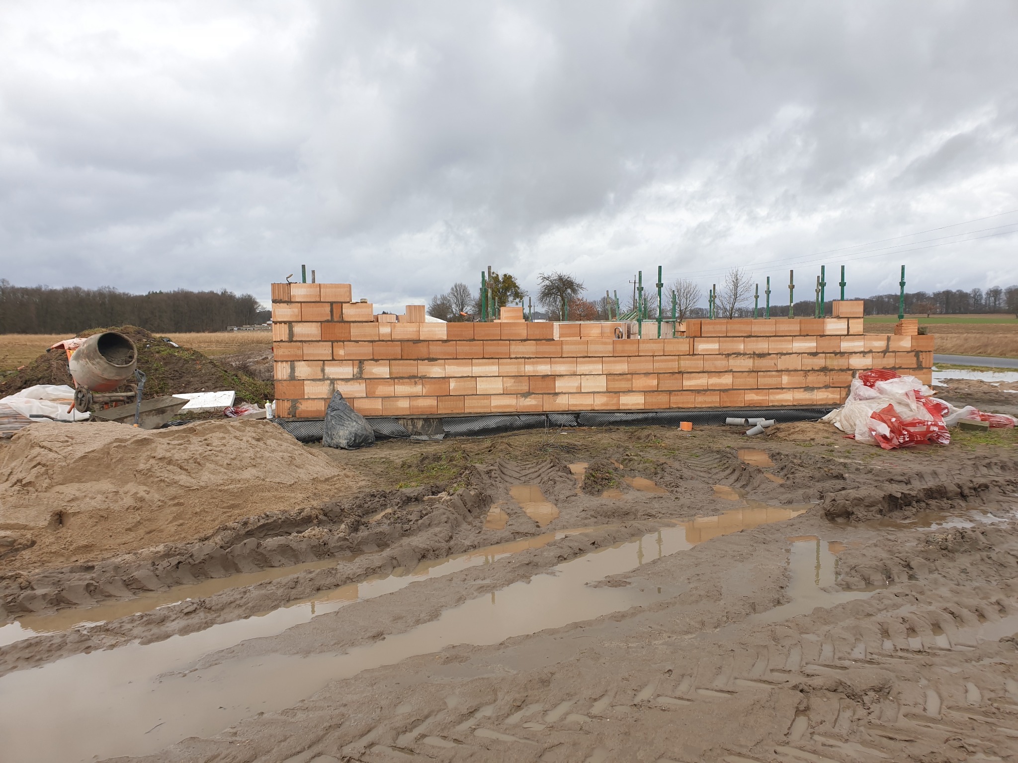 Wznoszenie muru z czerwonej cegły na budowie domu. Widoczne worki z zaprawą, betoniarka i błotniste podłoże. Pochmurne niebo w tle.