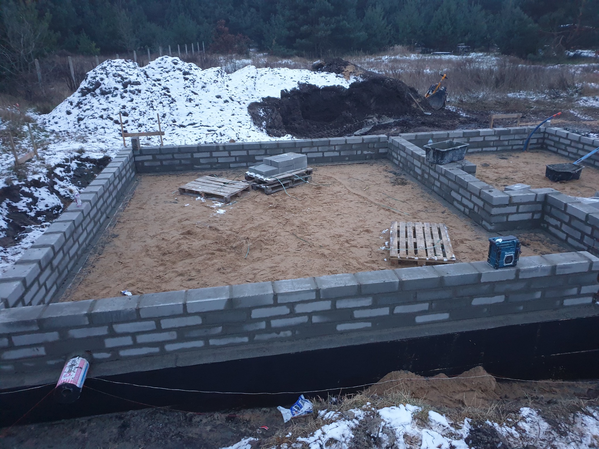 Fundamenty z bloczków betonowych w zimowej scenerii, częściowo ośnieżone. Widoczne palety, narzędzia, wykop zabezpieczony folią. Początek budowy domu.