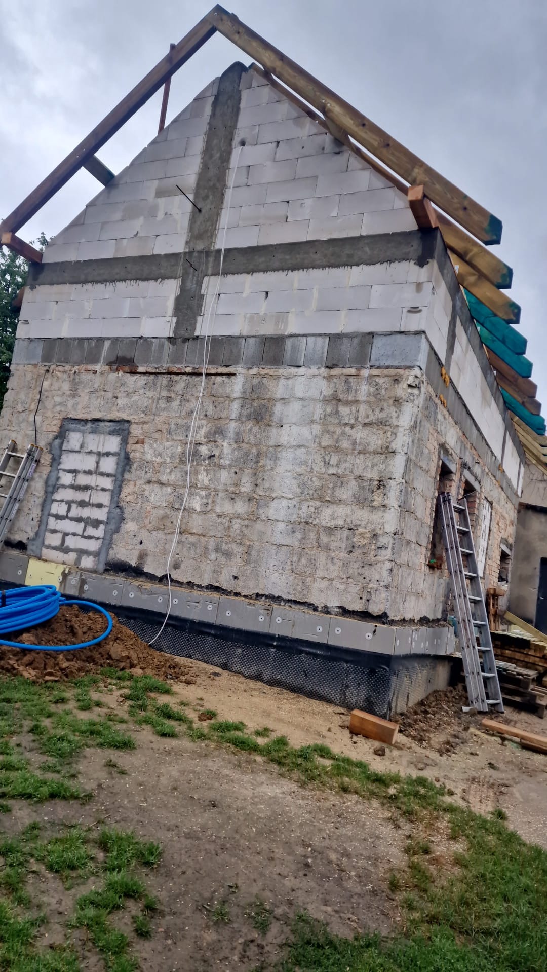 Budowa domu w trakcie: ściany z bloczków, częściowo wykonana więźba dachowa, widoczne materiały izolacyjne i drabiny. Prace murarskie na etapie wznoszenia ścian.