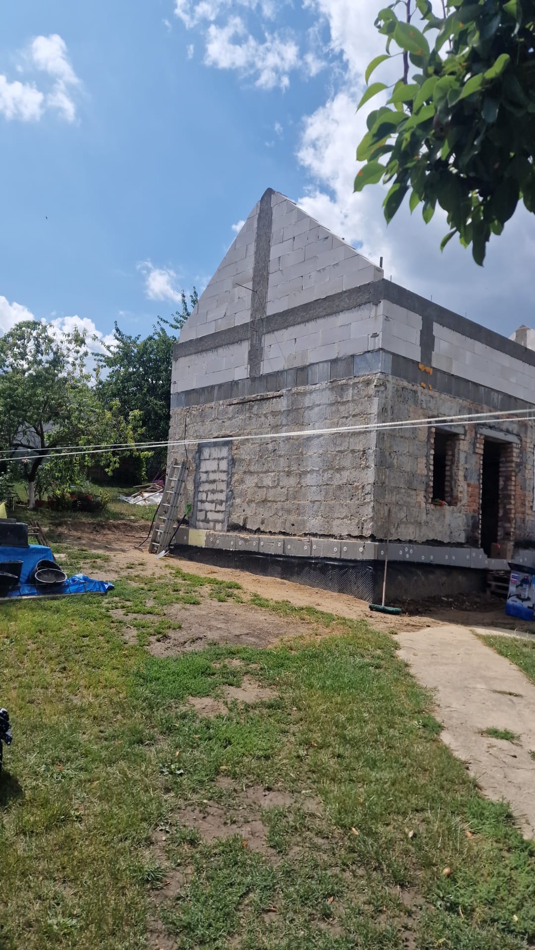 Budowa domu w toku: widoczne ściany z betonu komórkowego, częściowo otynkowane, z oknami i drzwiami. Na trawniku leżą narzędzia budowlane, oparta drabina. Błękitne niebo w tle.
