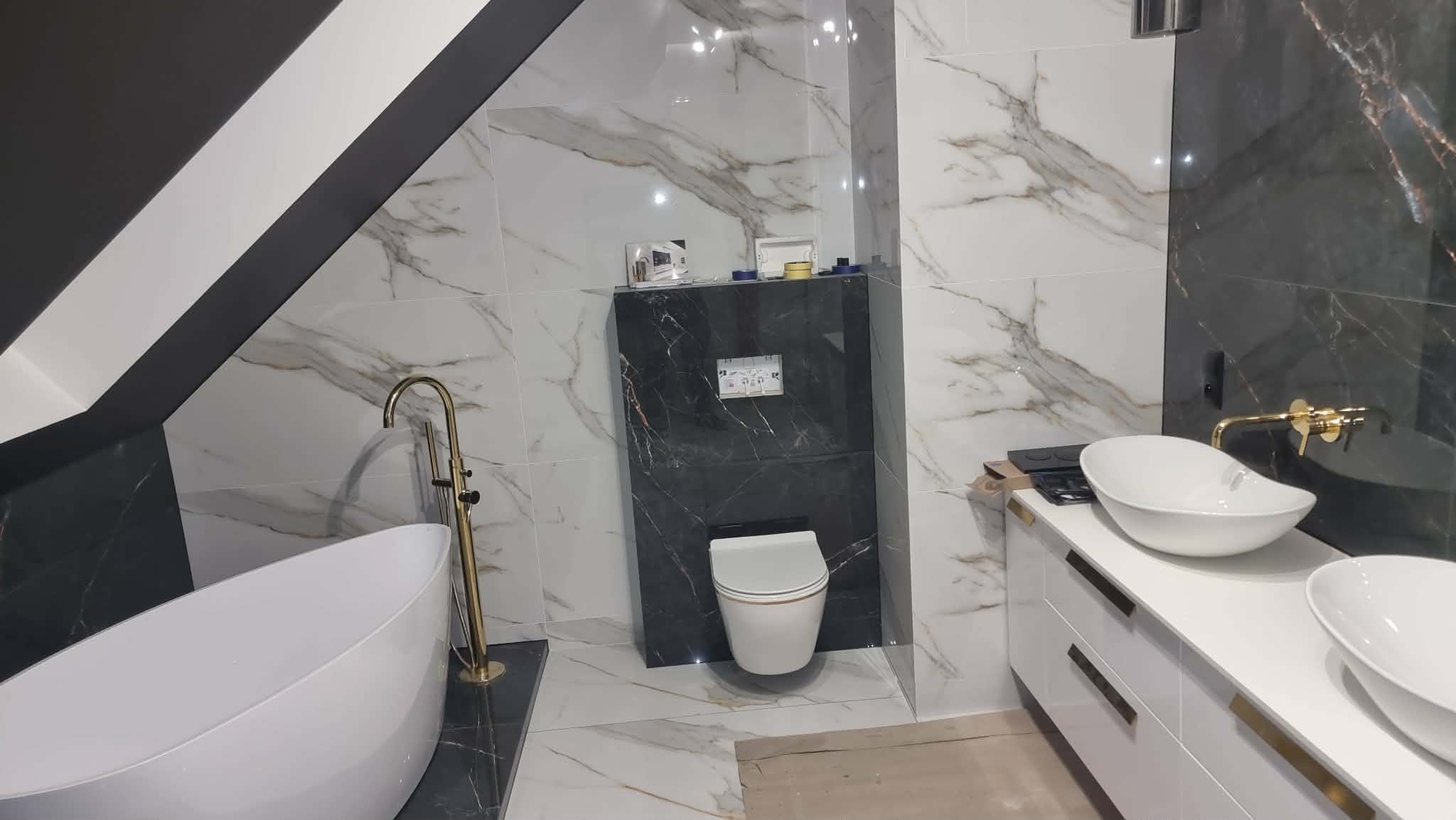 Elegancka łazienka w stylu glamour z marmurowymi płytkami, złotą armaturą i wolnostojącą wanną. Uwagę przykuwa kontrast czerni i bieli oraz skośny sufit.