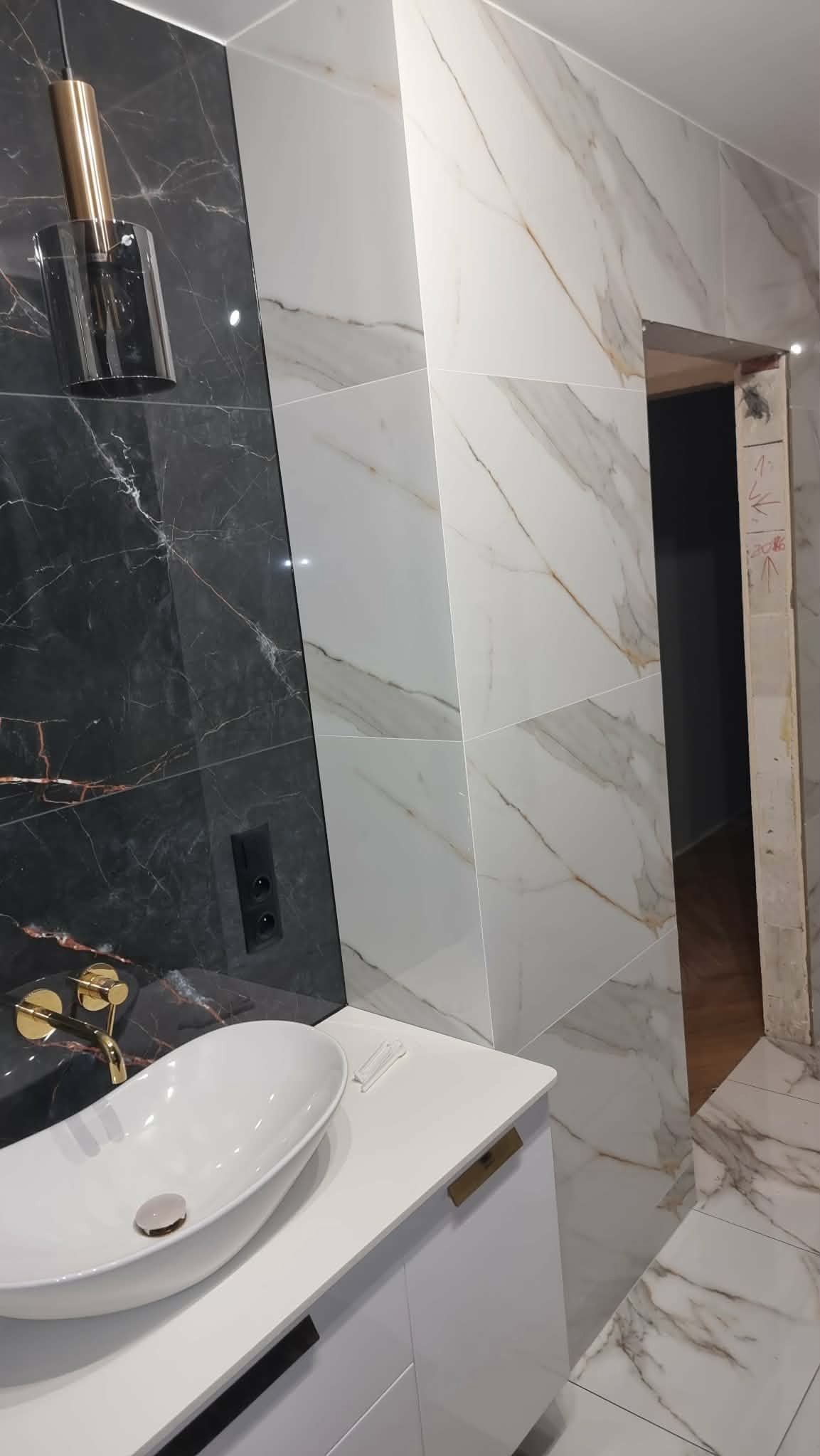 Wykończona łazienka z umywalką nablatową, złotą armaturą i marmurowymi płytkami w odcieniach bieli i czerni. Widoczny fragment drzwi bez ościeżnicy.