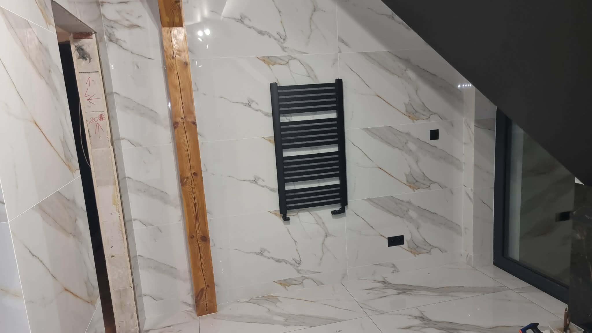 Wykończona łazienka na poddaszu z marmurowymi płytkami, czarnym grzejnikiem drabinkowym i oknem dachowym. Widoczna drewniana belka i fragment surowej ściany.