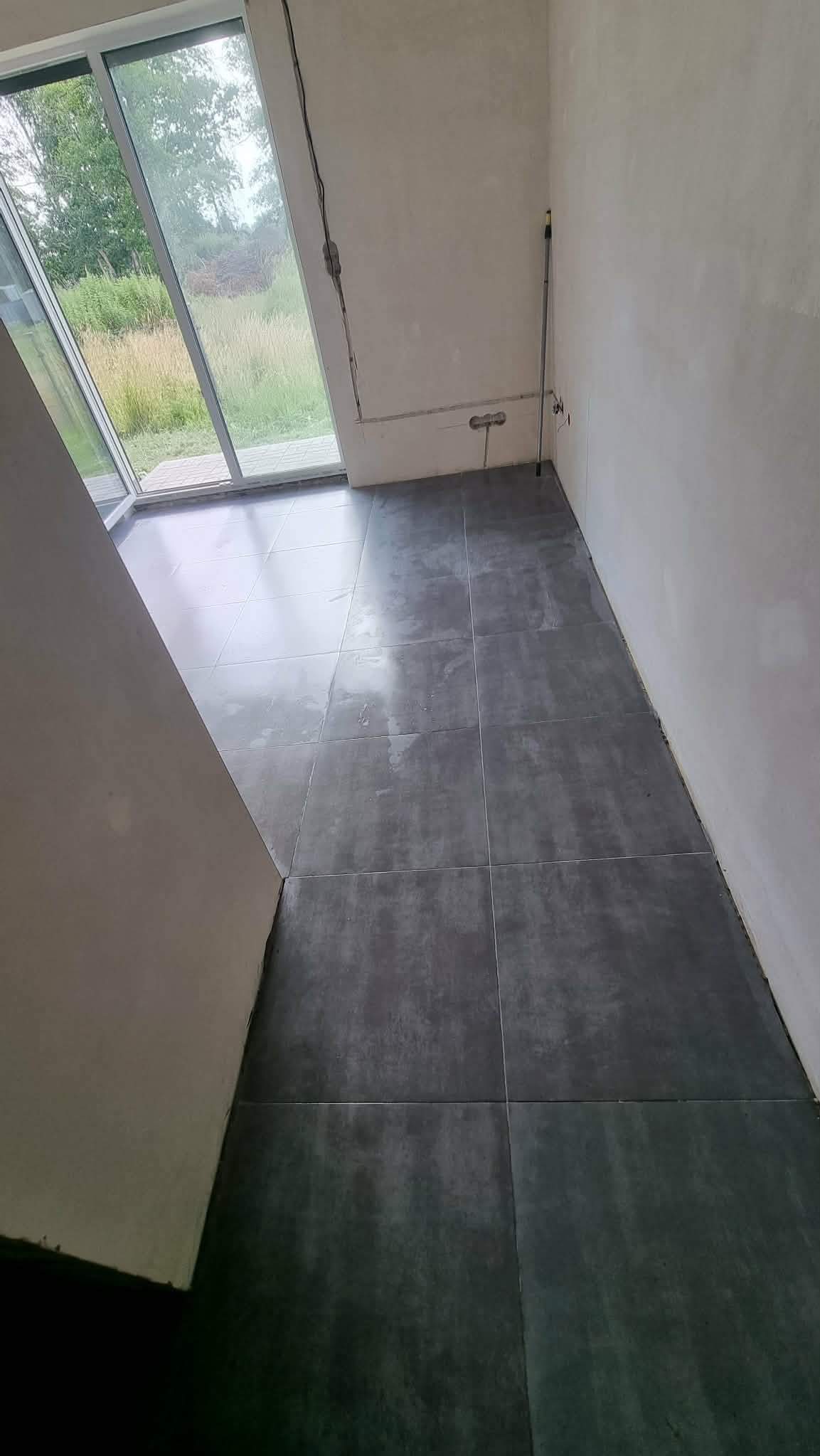 Ułożone, szare płytki podłogowe w surowym wnętrzu z dużym oknem tarasowym. Widoczne niedokończone ściany i wystające przewody.