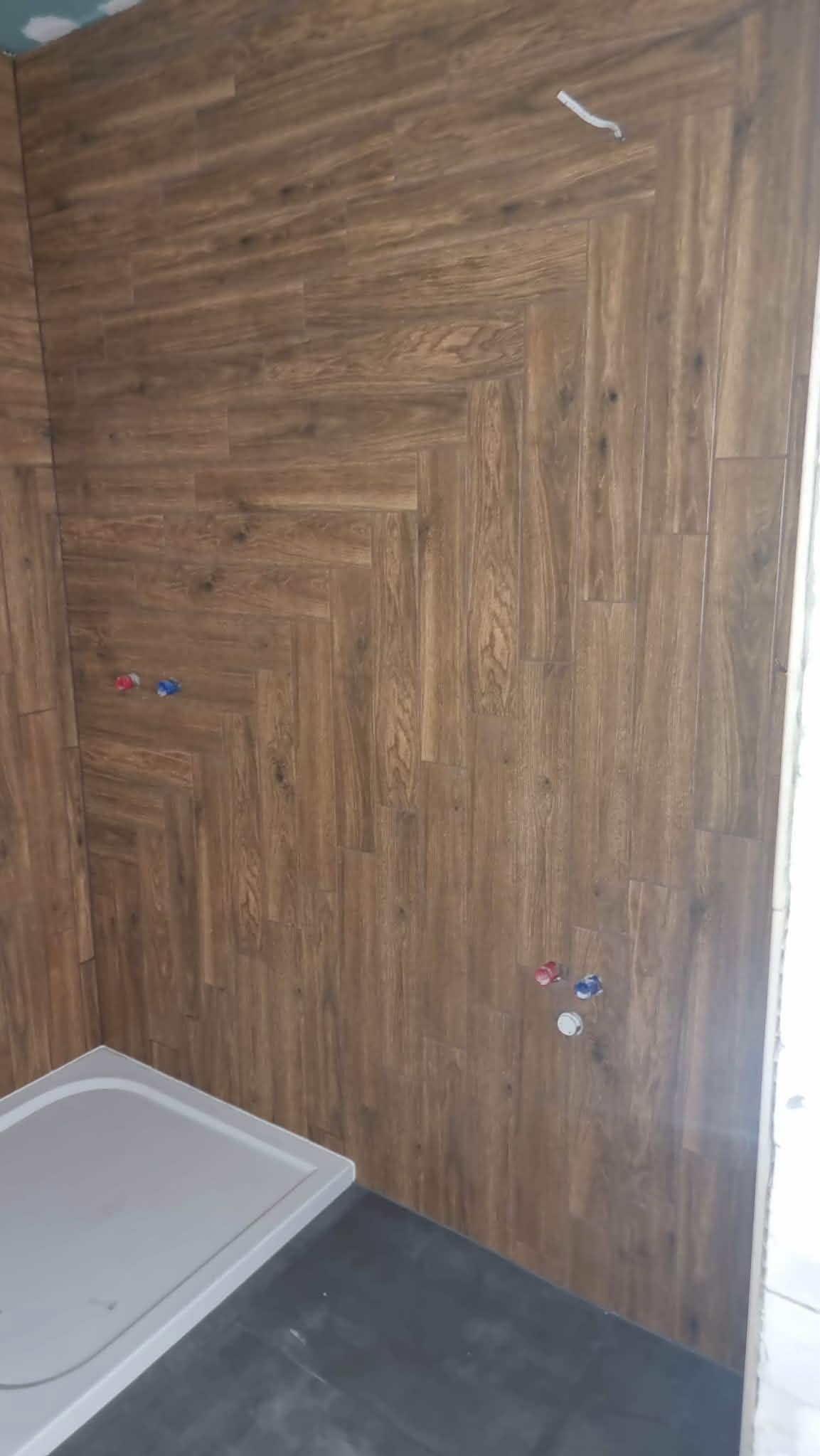 Wykończona łazienka z brązowymi płytkami imitującymi drewno ułożonymi w jodełkę na ścianie, widoczny brodzik i instalacja wodna.