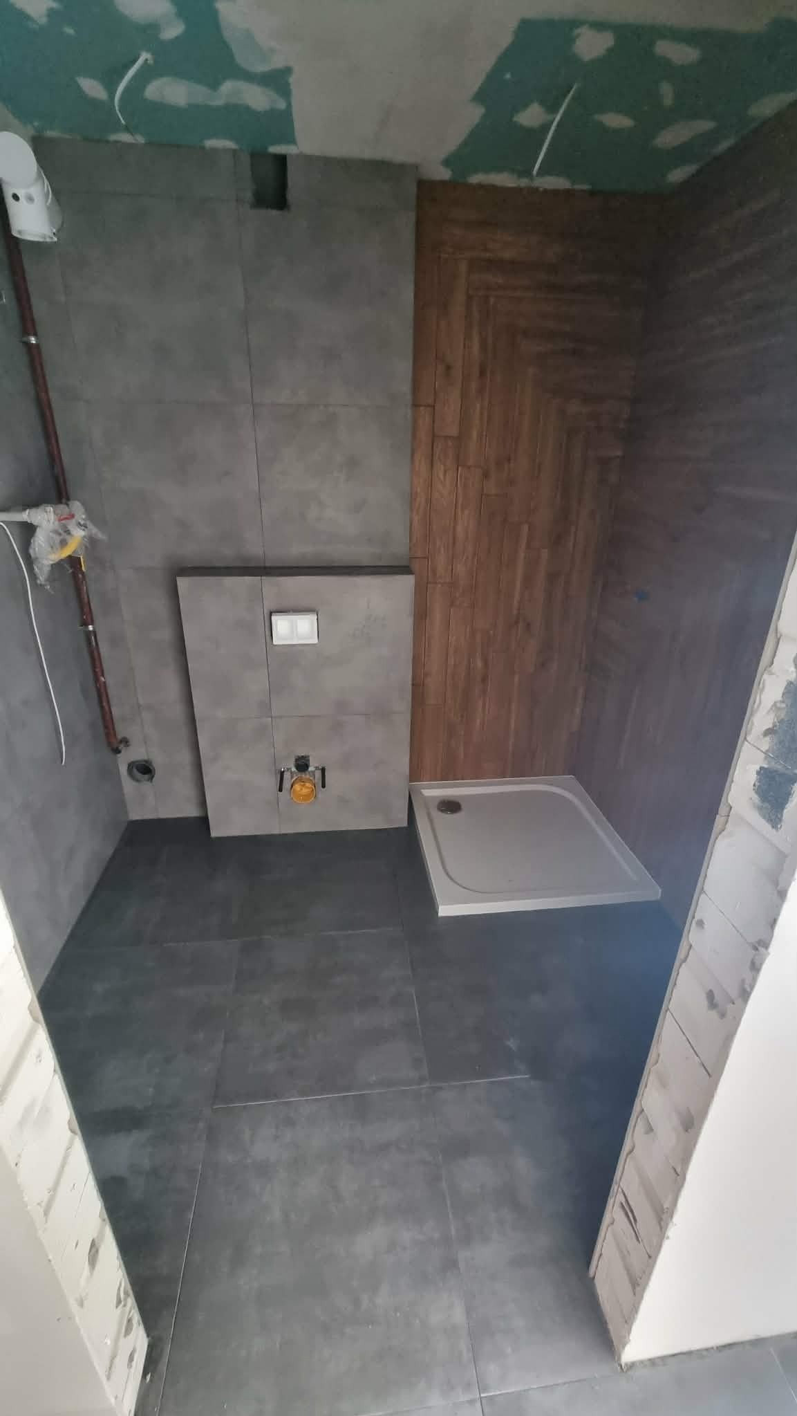 Wnętrze łazienki w trakcie remontu: szare płytki, drewnopodobna ściana, brodzik. Widoczne elementy instalacji wodno-kanalizacyjnej i niedokończone wykończenie sufitu.