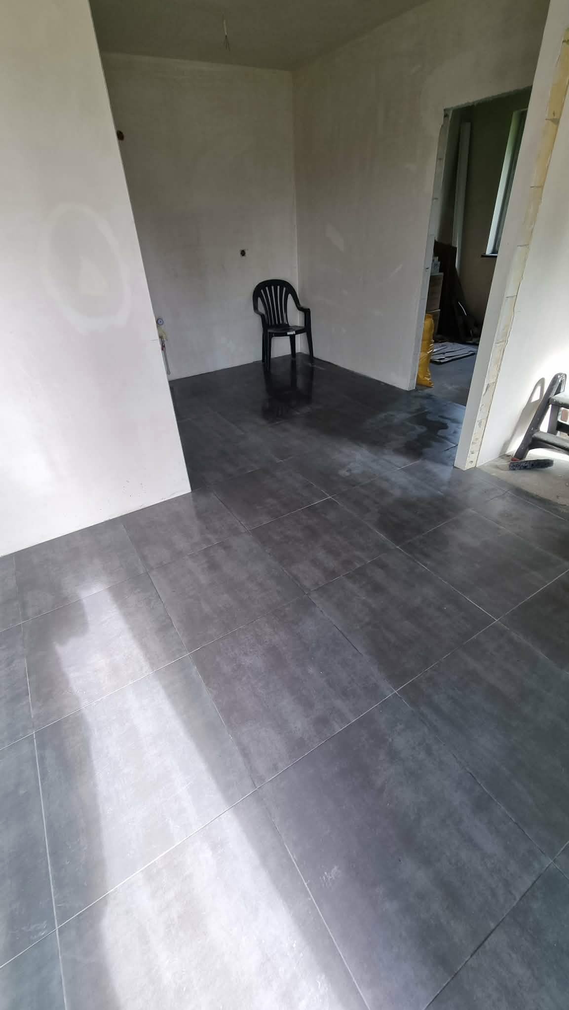 Ułożone, ciemnoszare płytki podłogowe w surowym wnętrzu z czarnym krzesłem w tle. Widoczny fragment drabiny malarskiej.