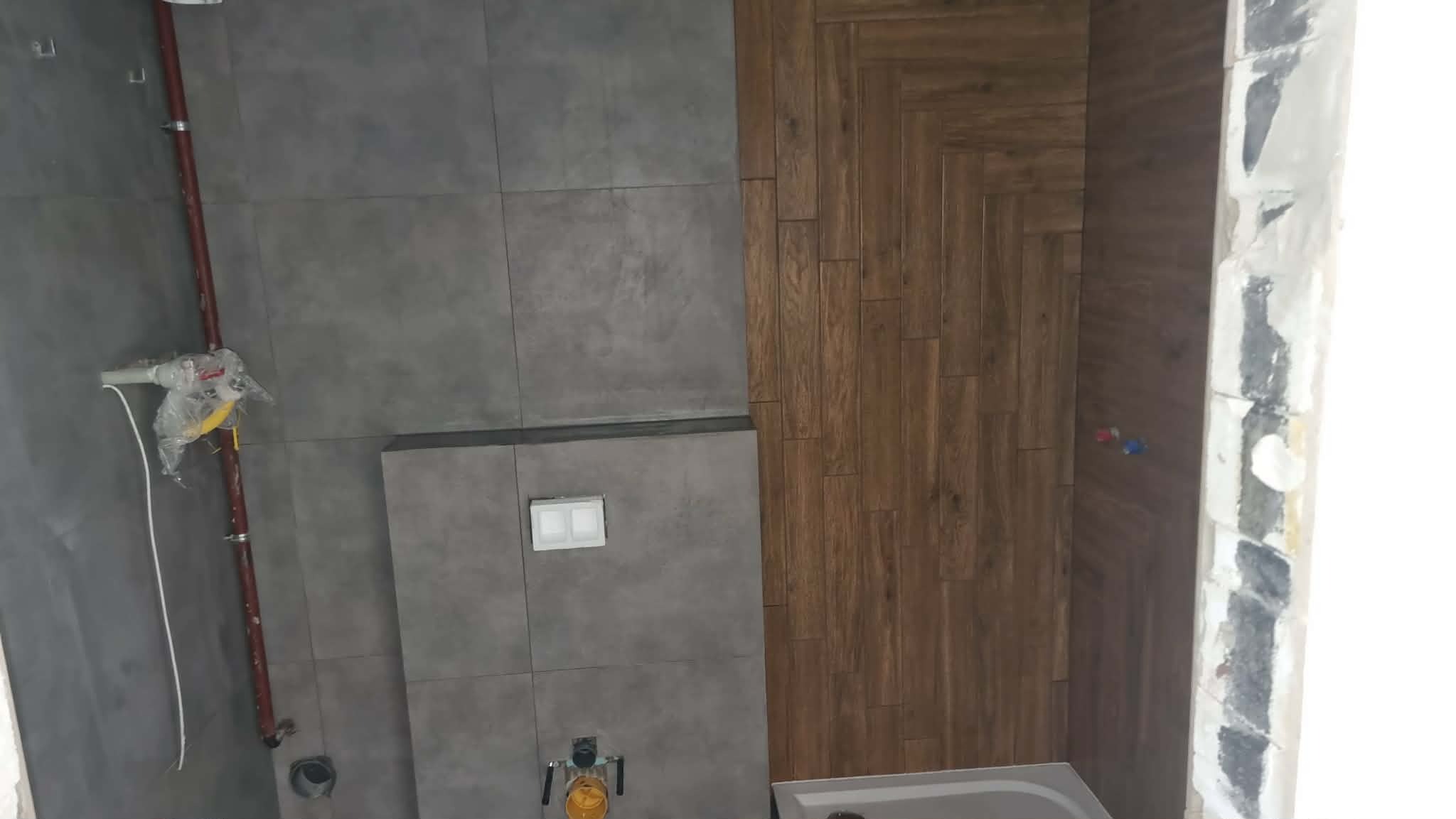 Szare płytki ścienne i drewnopodobne ułożone w jodełkę w łazience w trakcie remontu. Widoczne rury i instalacje. Częściowo widoczna wanna.