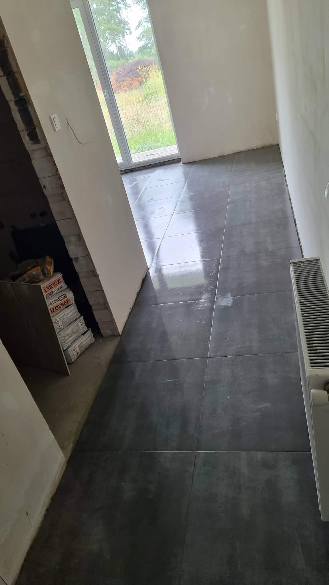 Ułożona, ciemna podłoga z płytek w surowym wnętrzu. Widoczne worki z zaprawą i grzejnik. Przez okno widać zieleń.