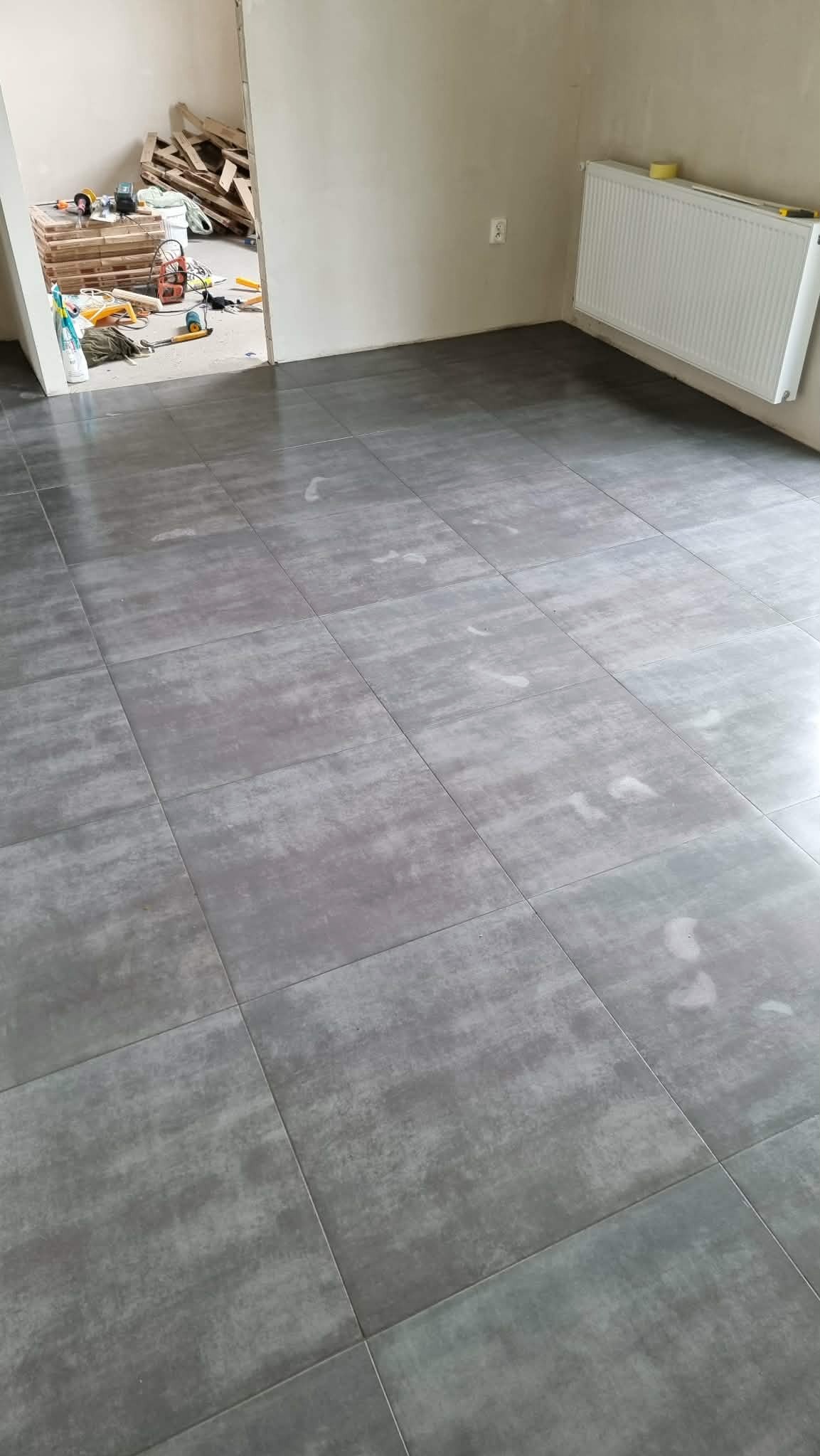 Ułożona, szara, matowa podłoga z płytek w nowym pomieszczeniu. W tle widoczne narzędzia i materiały budowlane, grzejnik na ścianie.