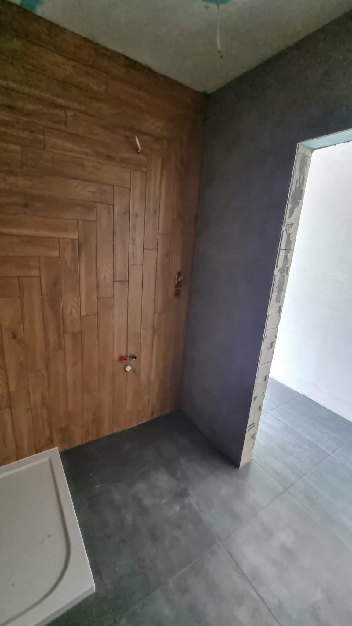 Wnętrze łazienki w trakcie wykańczania: drewnopodobna okładzina ścienna ułożona w jodełkę, szara ściana i podłoga z płytek, widoczne przyłącza wodne.