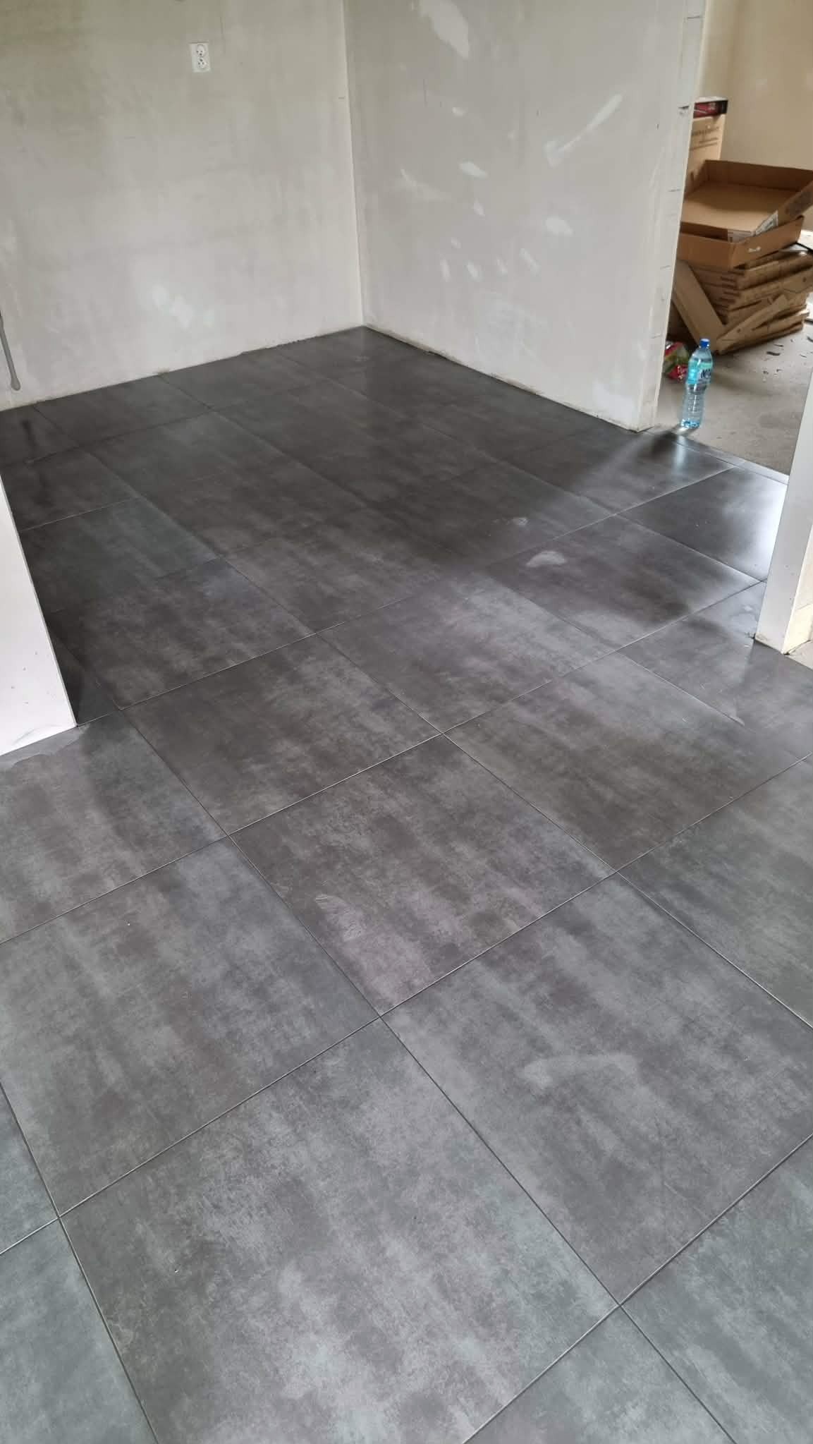 Ułożone, szare płytki podłogowe w surowym wnętrzu. Widoczne kartony i butelka wody w tle. Minimalistyczny design z efektem betonu.