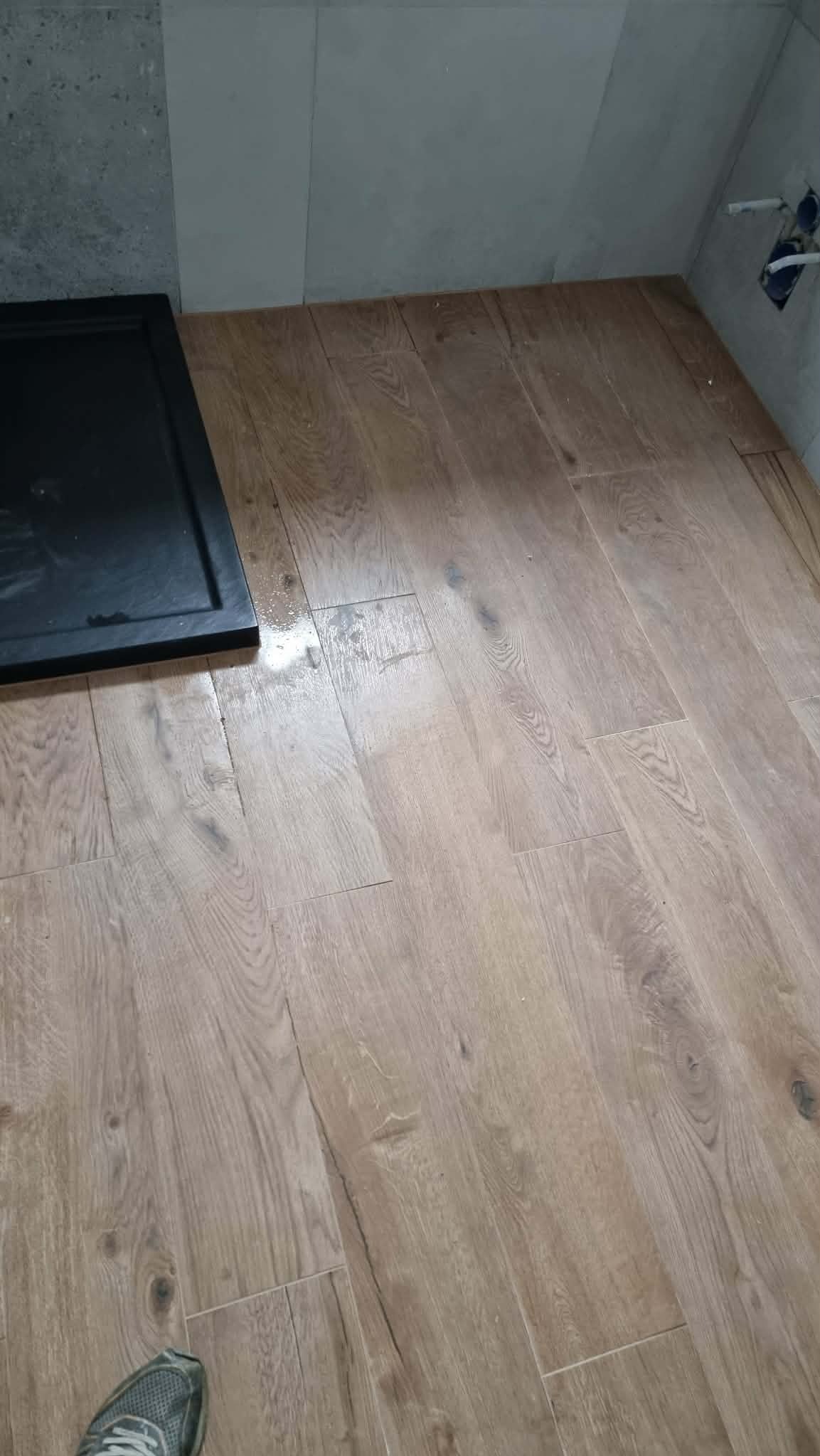 Ułożona podłoga z paneli imitujących drewno w łazience, widoczny brodzik i fragment buta w lewym dolnym rogu. Szare płytki na ścianie.