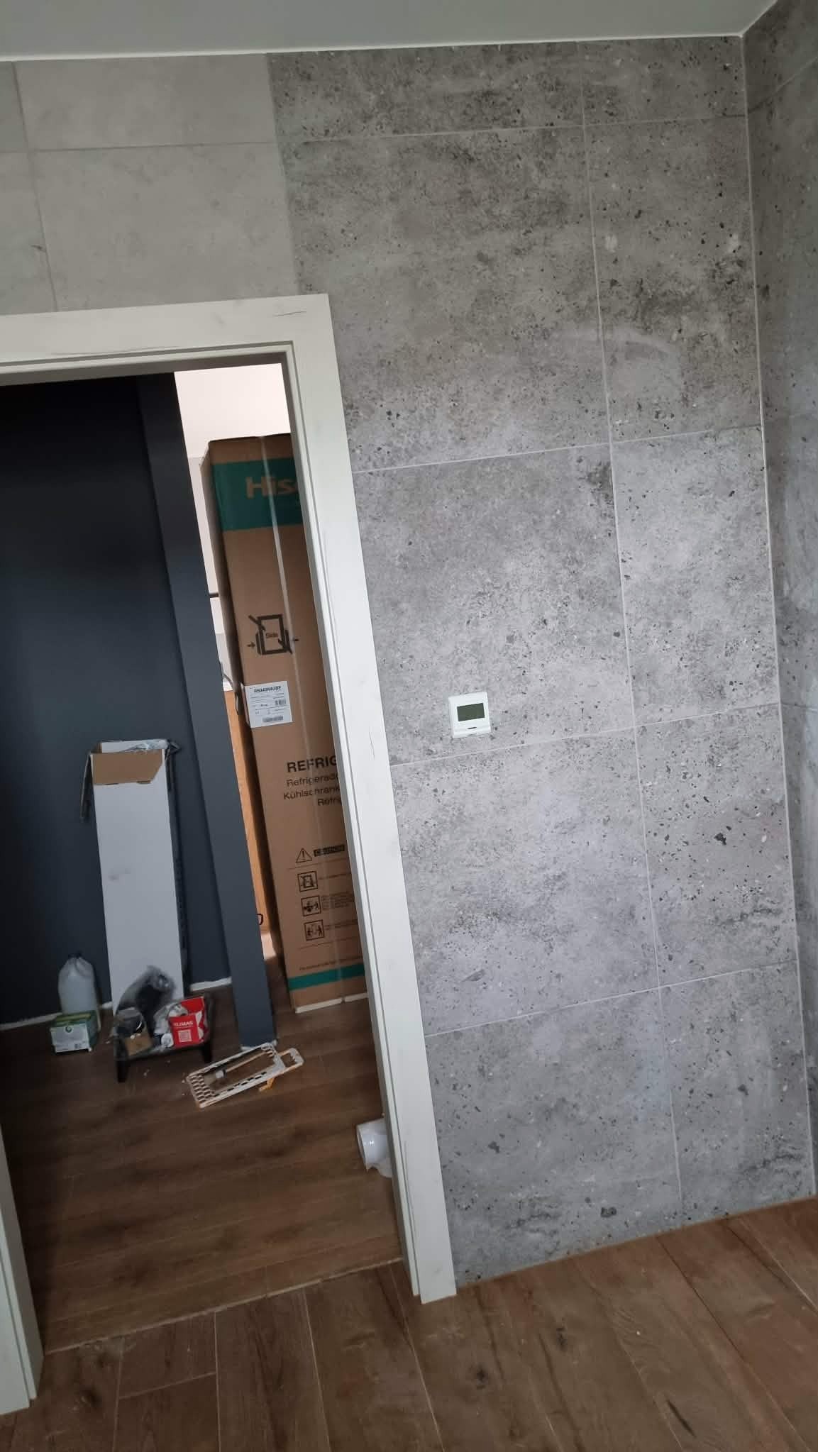 Wykończona ściana z szarych płytek imitujących kamień, widoczny fragment futryny drzwi i kartonowe opakowanie w tle. Minimalistyczny termostat na ścianie.