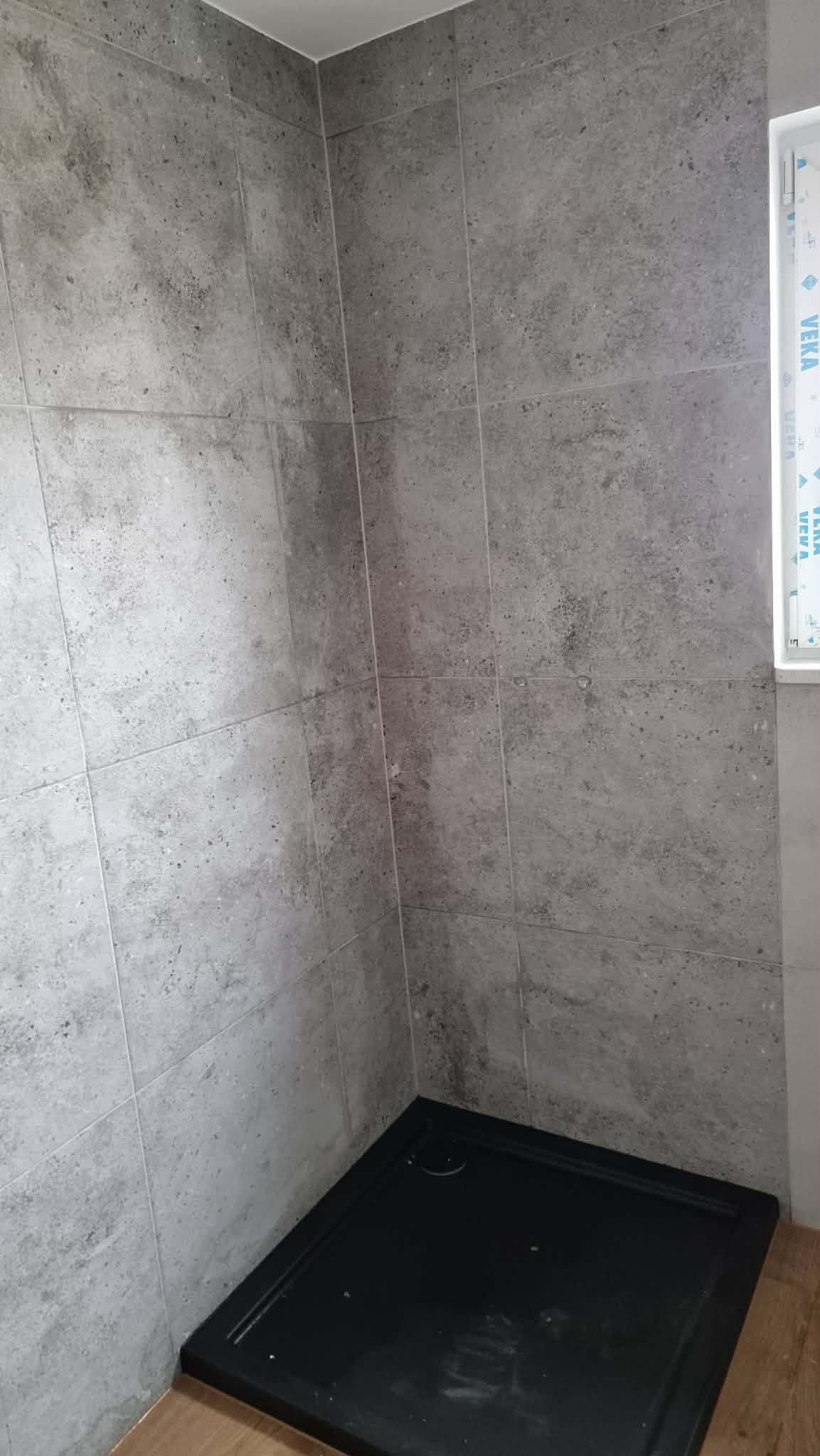 Wykończona kabina prysznicowa z szarymi płytkami imitującymi kamień i czarnym brodzikiem. Widoczne okno z profilem PCV. Minimalistyczny design.