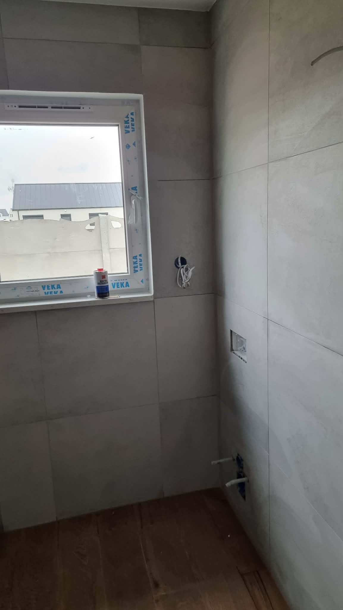 Wnętrze łazienki w trakcie wykańczania: szare płytki, okno z widokiem na dom, instalacja wodna i elektryczna w ścianie, drewniana podłoga. Widoczny klej do okien.