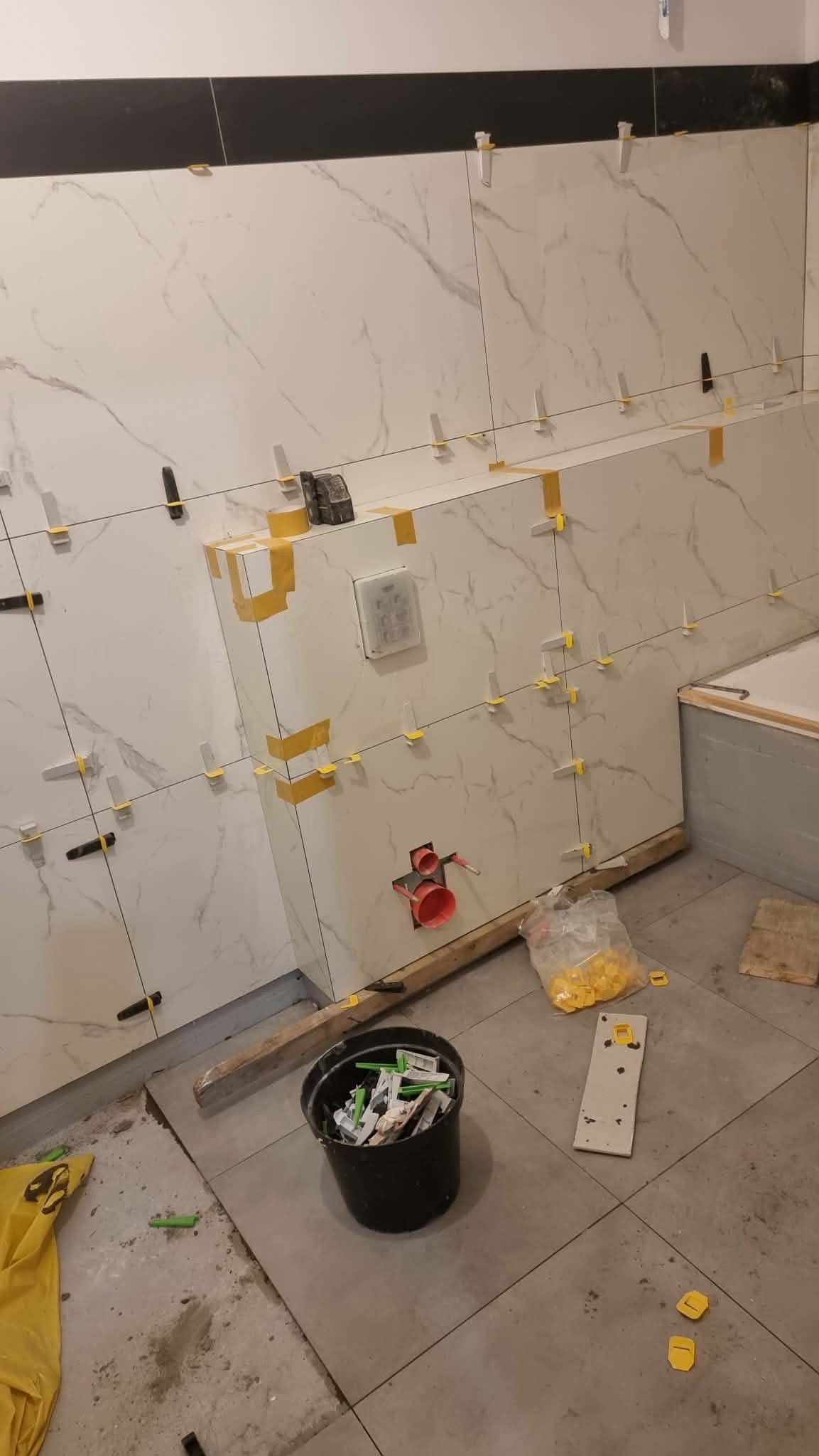 Układanie białych płytek imitujących marmur na ścianie z użyciem poziomowania. Widoczne narzędzia i akcesoria glazurnicze, w tym wiadro z krzyżykami dystansowymi.