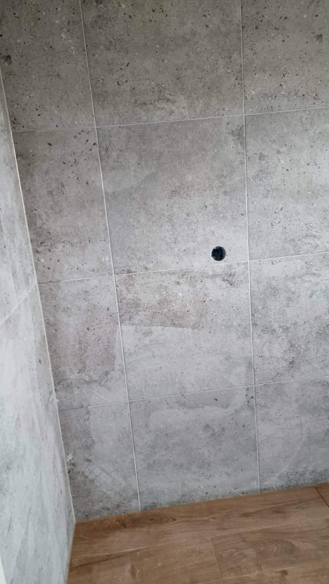 Szare, kwadratowe płytki ścienne z fugami i otworem na instalację, dolna krawędź styka się z panelami podłogowymi imitującymi drewno.