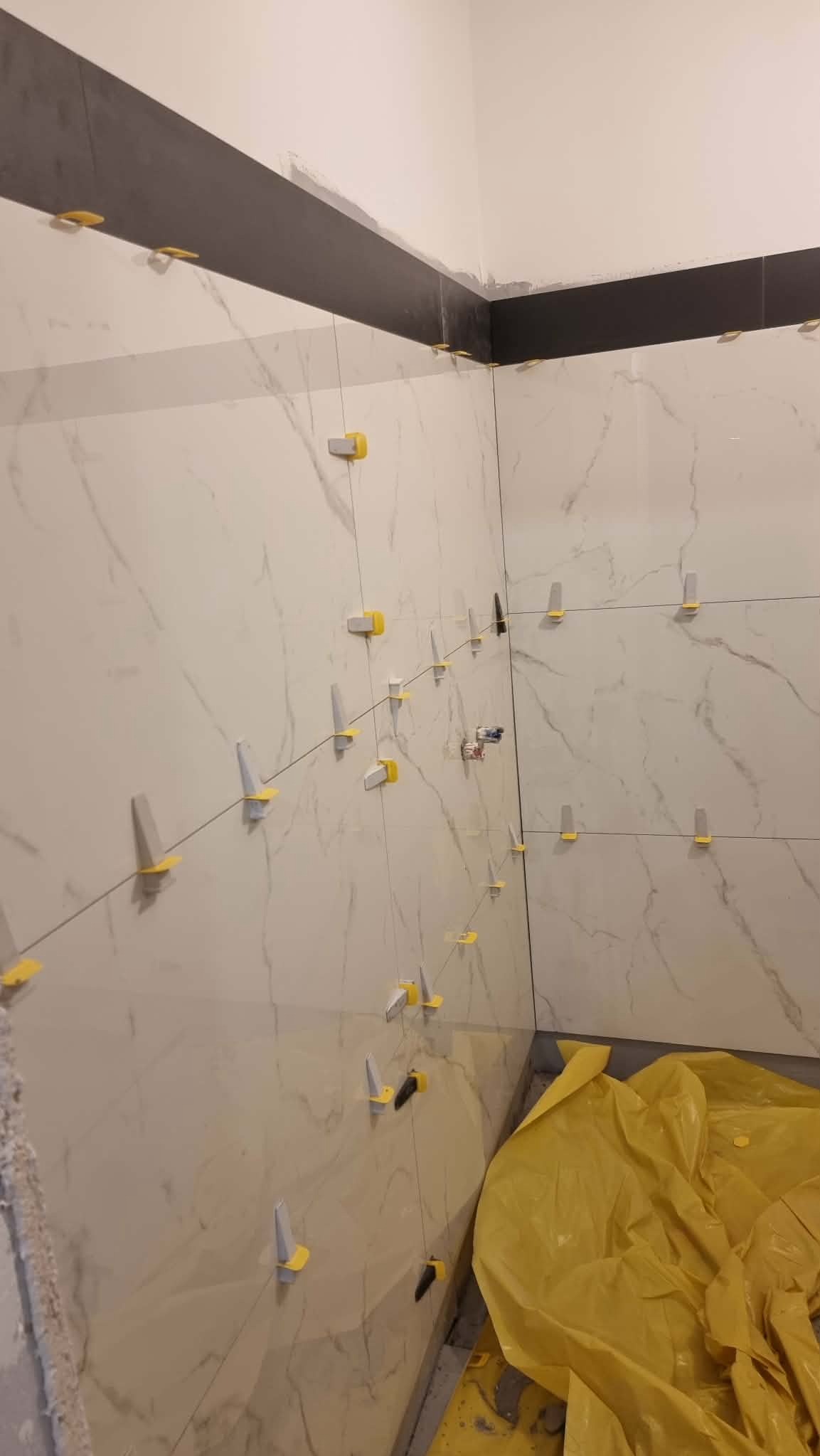 Układanie płytek imitujących marmur na ścianie z użyciem systemu poziomowania. Widoczne żółte klipsy i czarna listwa dekoracyjna u góry. Podłoga zabezpieczona folią.