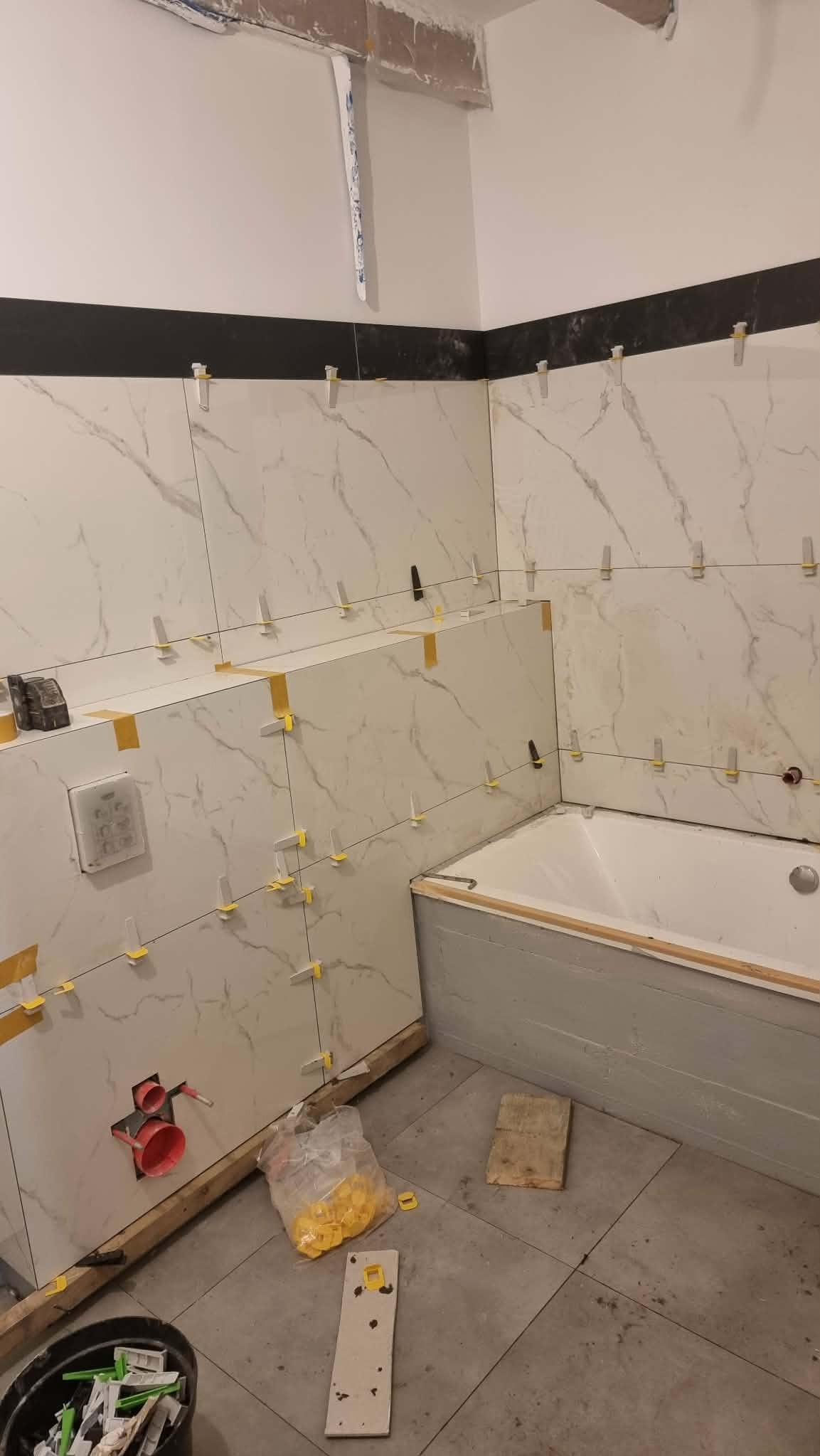 Układanie białych płytek imitujących marmur w łazience, widoczne krzyżyki dystansowe i poziomice. Etap prac wykończeniowych, przed fugowaniem.