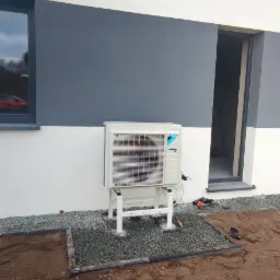 Zewnętrzna jednostka klimatyzacji Daikin zamontowana na białym stelażu, z widocznymi przewodami, na tle szaro-białej ściany budynku i grządką z szarych kamieni.