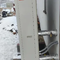 Zewnętrzna jednostka pompy ciepła Daikin zamontowana na betonowym fundamencie przy ścianie budynku w zimowej scenerii, z widocznymi izolowanymi rurami i zaworami.