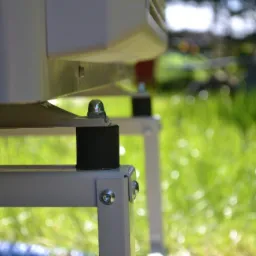 Fragment konstrukcji wsporczej urządzenia z widocznymi elementami mocującymi (śruby, nakrętki) i gumowymi podkładkami antywibracyjnymi, na tle rozmytej zieleni trawnika.