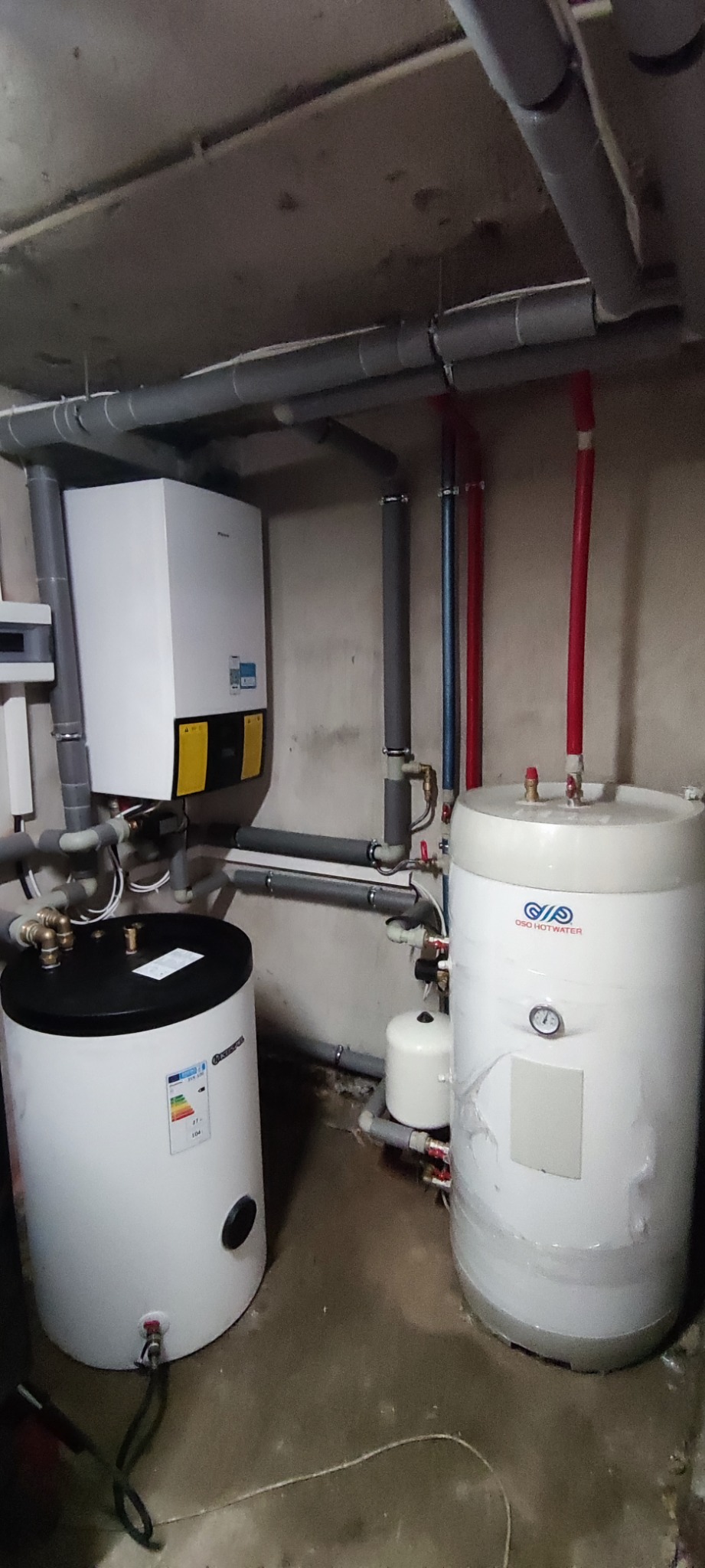 Zamontowany system grzewczy z dwoma zasobnikami ciepłej wody OSO HOTWATER, piecem CO z żółtymi etykietami i szarymi rurami w surowym pomieszczeniu.
