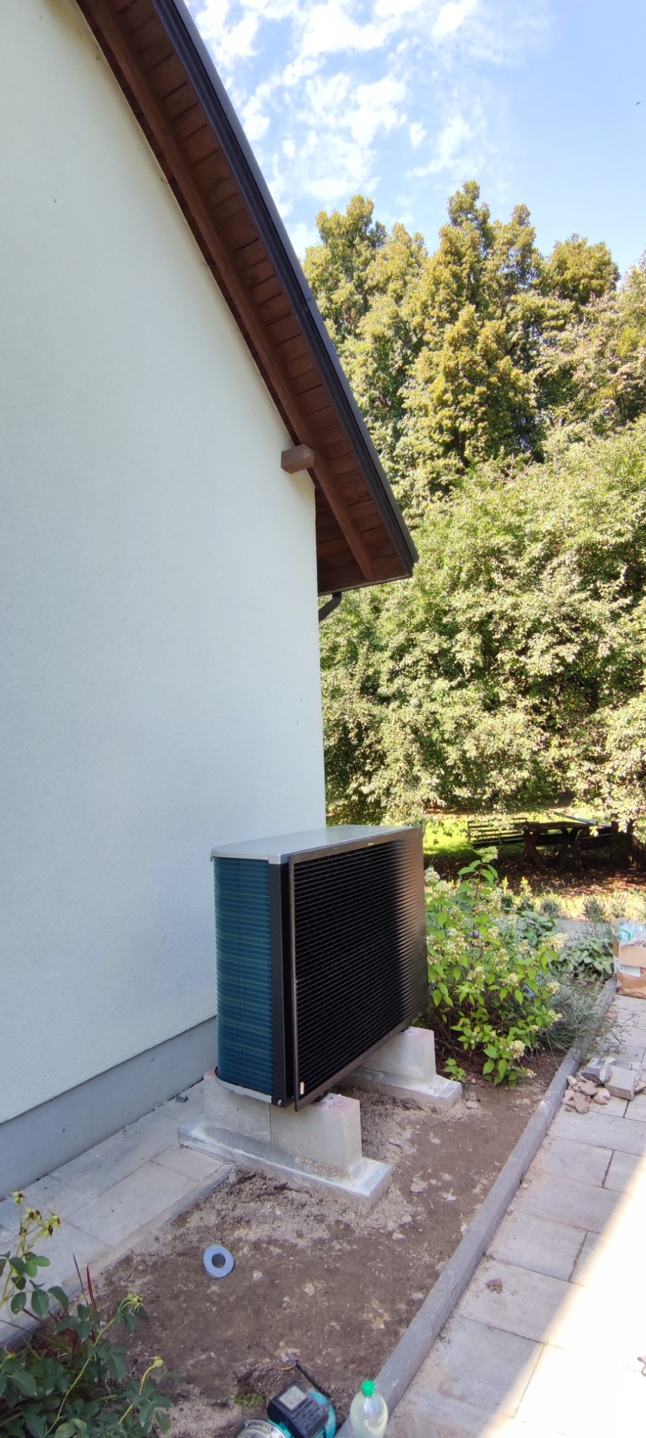 Zewnętrzna jednostka pompy ciepła ustawiona na betonowych wspornikach przy ścianie budynku, widoczne elementy instalacji, kostka brukowa i zieleń w tle.