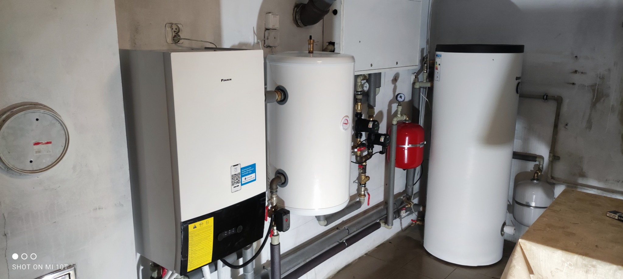 Zestaw urządzeń grzewczych: biały piec Daikin, dwa białe zbiorniki, czerwony zbiornik przeponowy, rury i armatura w surowym pomieszczeniu.