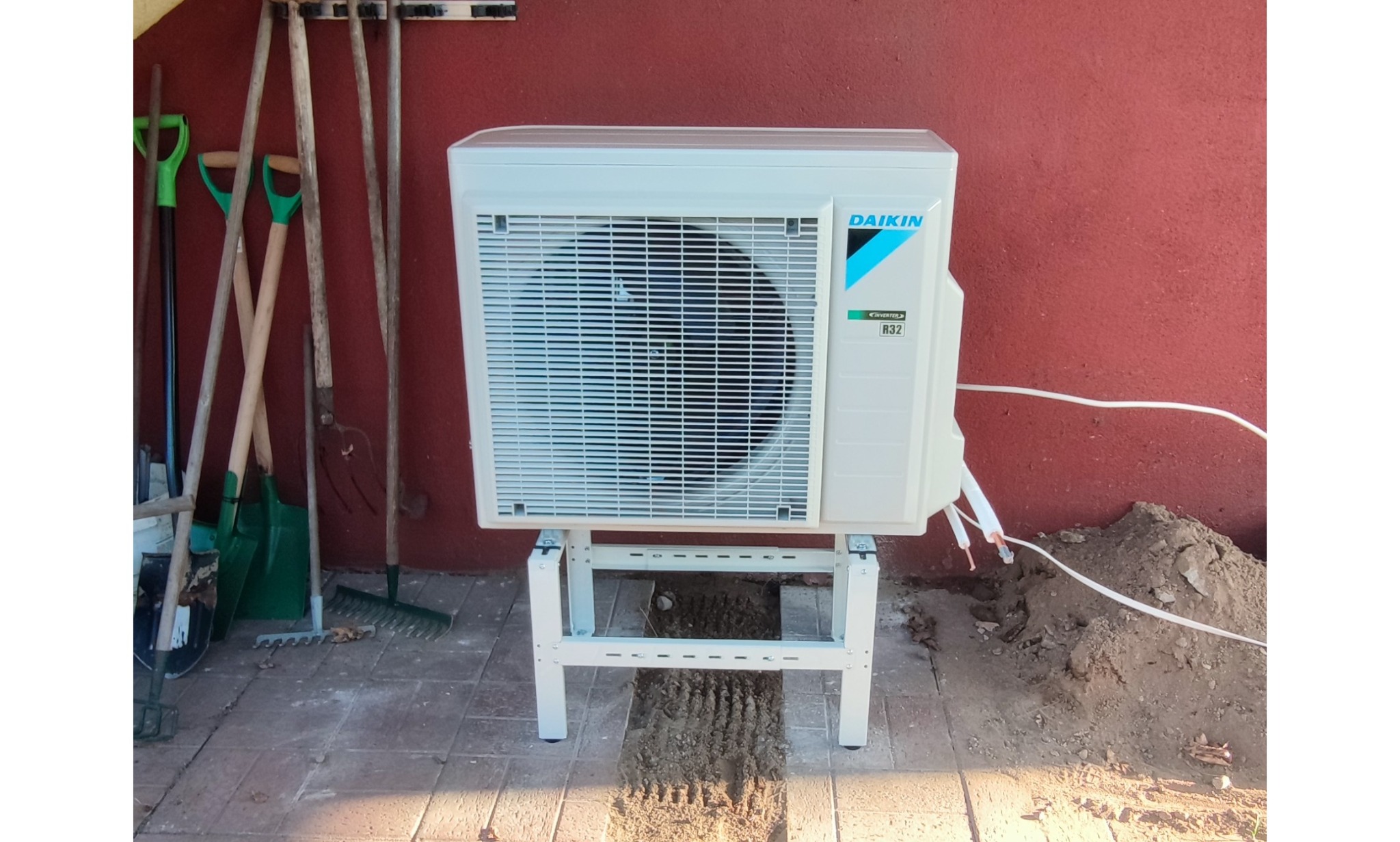 Biała jednostka zewnętrzna klimatyzatora Daikin zamontowana na metalowym stojaku, z widocznymi przewodami i stertą ziemi obok, na tle czerwonej ściany i narzędzi ogrodowych.