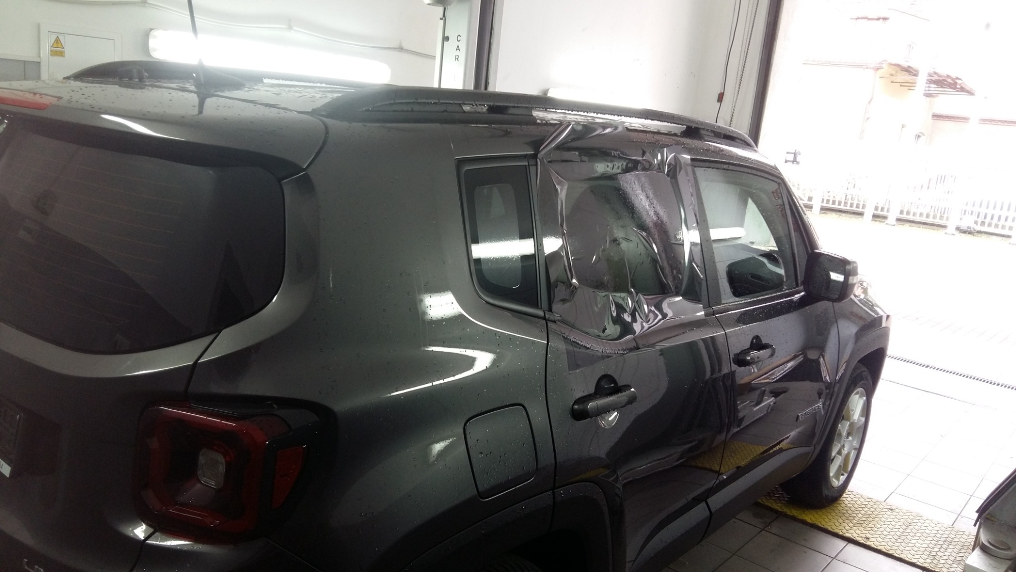 Szary Jeep Renegade w trakcie aplikacji folii na tylnych bocznych szybach, widoczne wstępnie docięte arkusze folii na oknach, wnętrze warsztatu z jasnym oświetleniem.