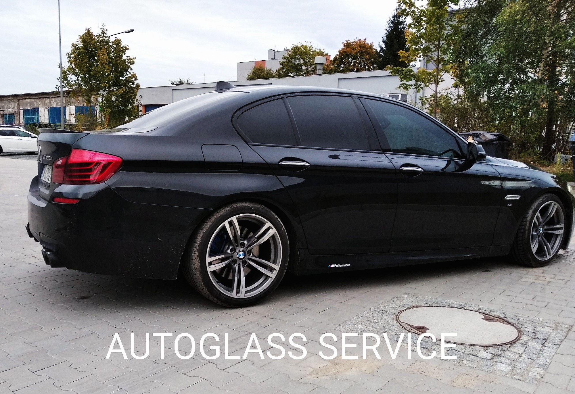 Czarne BMW serii 5 z przyciemnionymi szybami bocznymi i tylną, zaparkowane na betonowym placu, widok z tyłu na bok, z widocznym logo M Performance.