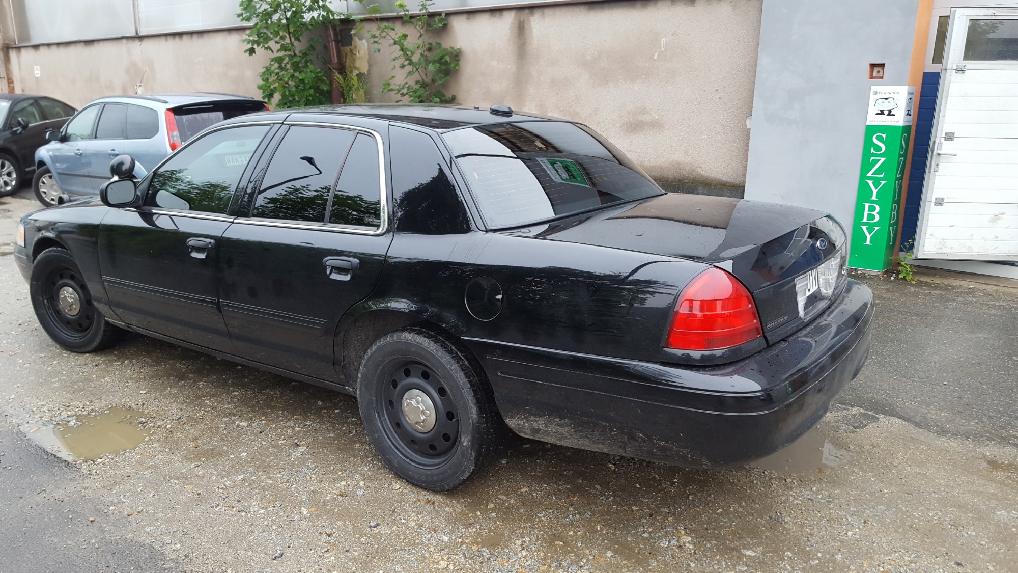 Czarny samochód Ford Crown Victoria z przyciemnionymi szybami, zaparkowany na żwirowym podłożu z kałużami, widoczny z tyłu i boku. Na tylnej szybie widoczne poziome paski folii. Obok samochodu...