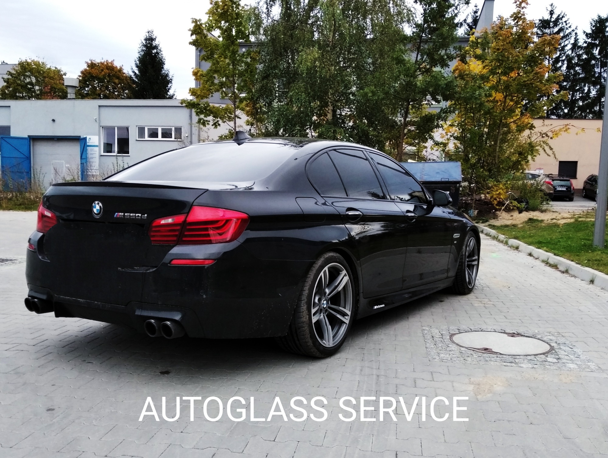 Czarne BMW serii 5 po zabiegu przyciemniania szyb, widoczne ciemne szyby boczne i tylna, na tle budynku i drzew, z napisem 'AUTOGLASS SERVICE' na dole zdjęcia.